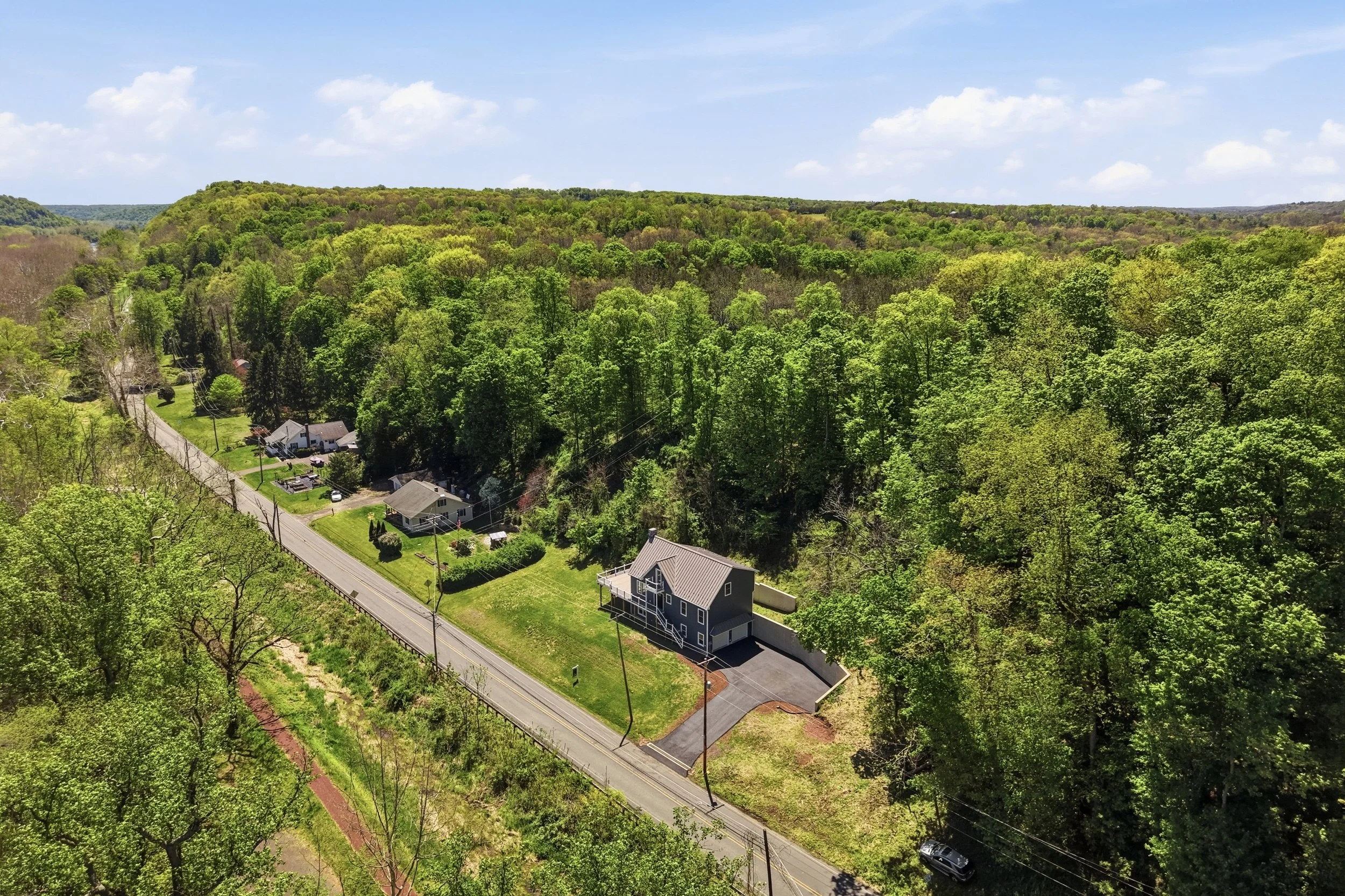Aerial view of a residential area at the edge of a dense, green forest under a partly cloudy sky. Houses with yards are situated along a main road, with power lines running through the neighborhood.