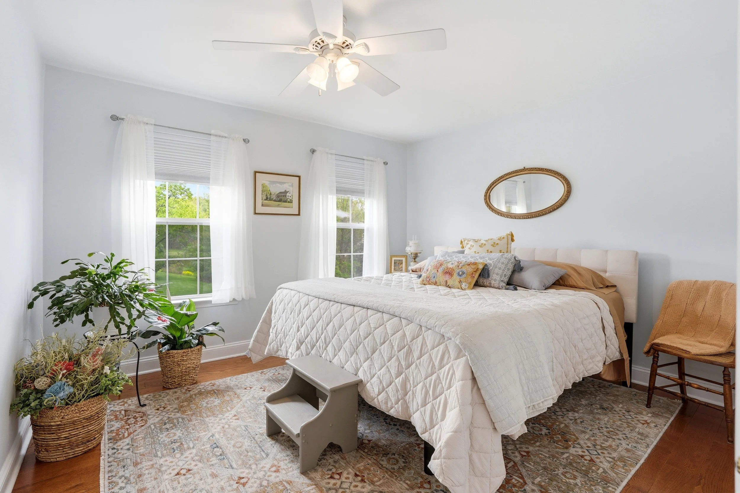 A bedroom with two large windows, white walls, a ceiling fan with lights, a bed with a white quilt and multiple pillows, a vintage mirror on the wall, a wicker chair, potted plants, and a framed picture of a house outside the window.
