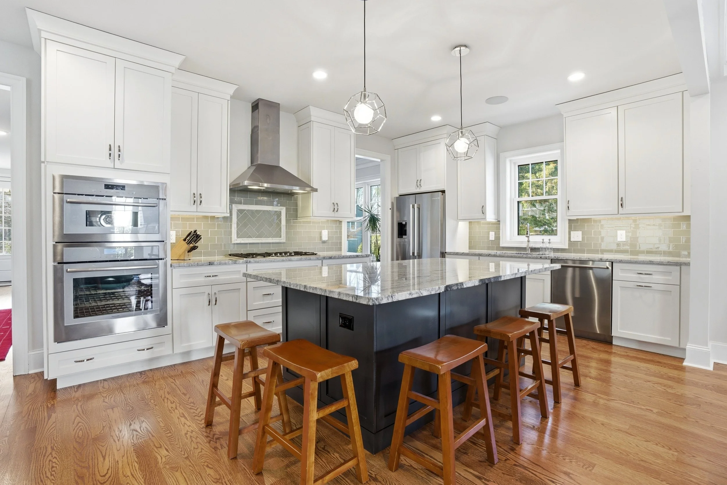 A modern kitchen with white cabinets, stainless steel appliances, a gray tile backsplash, a large island with a granite countertop, and wooden stools. Double pendant lights hang over the island. There are windows with natural light and hardwood floor