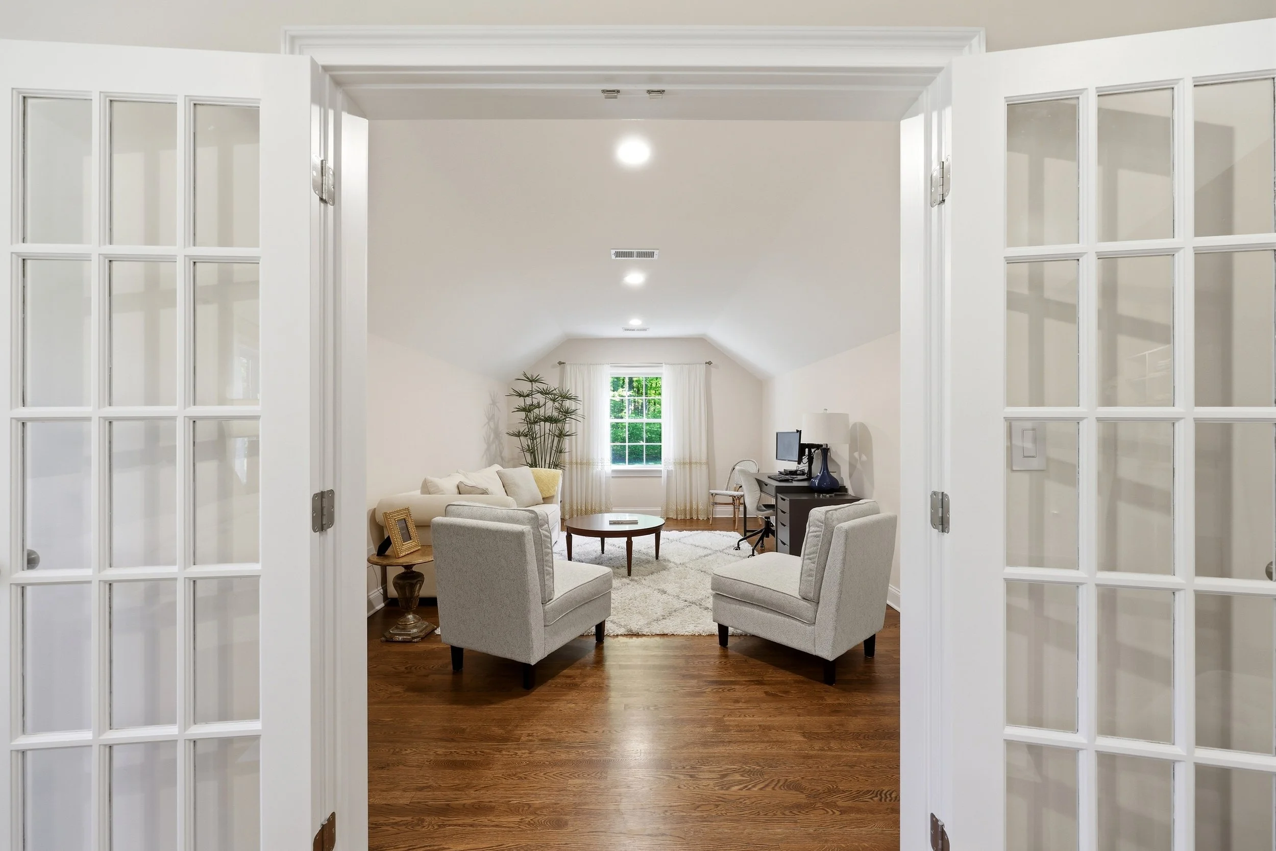 Looking through double glass doors into a bright, modern living room with hardwood floors, white walls, a white sofa, armchairs, a small round wooden table, a window with curtains, a tall plant, and a desk with a computer and lamp.