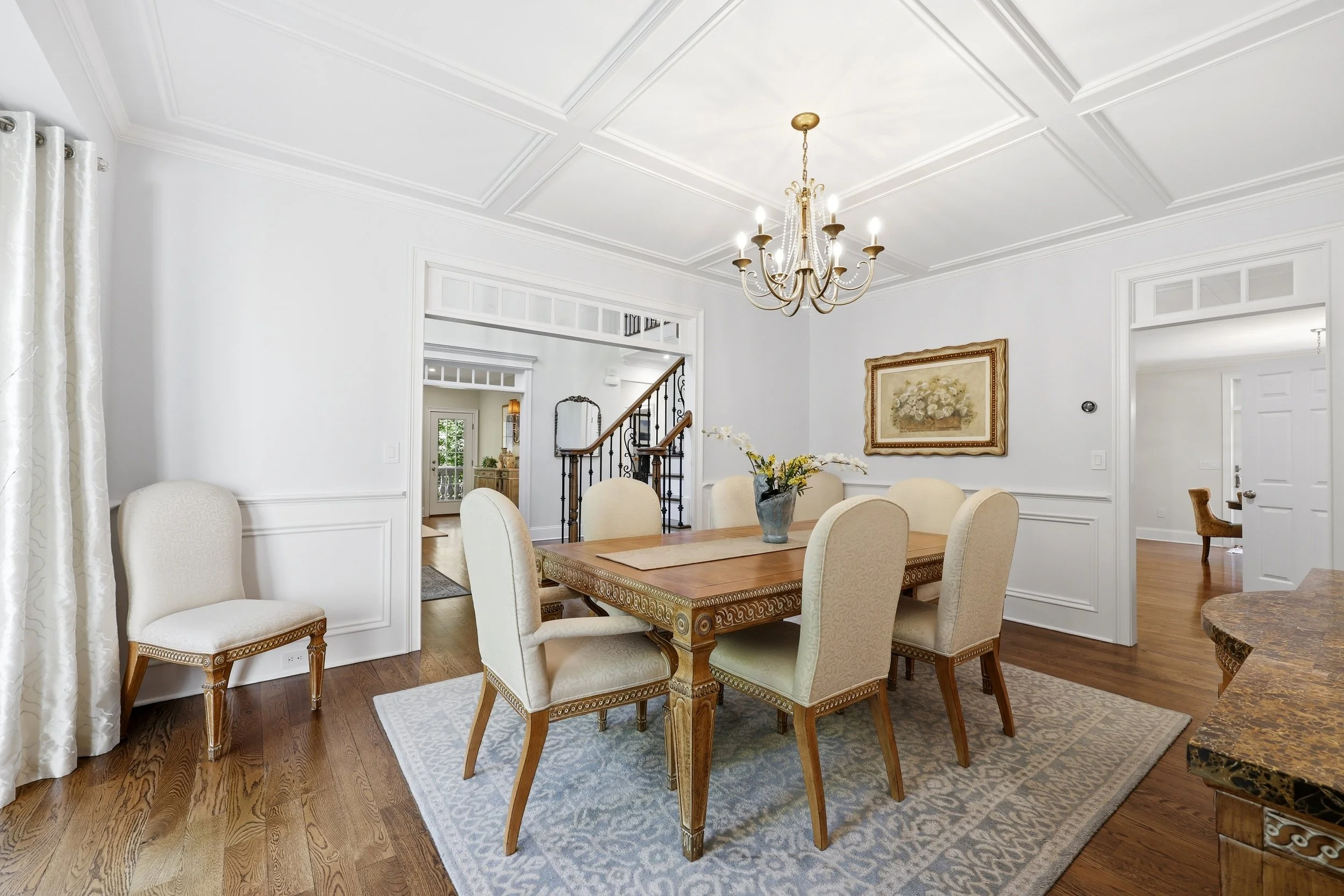 A dining room with a wooden table surrounded by cream-colored upholstered chairs, a chandelier hanging from the ceiling, a framed floral painting on the wall, and hardwood floors with a patterned rug.