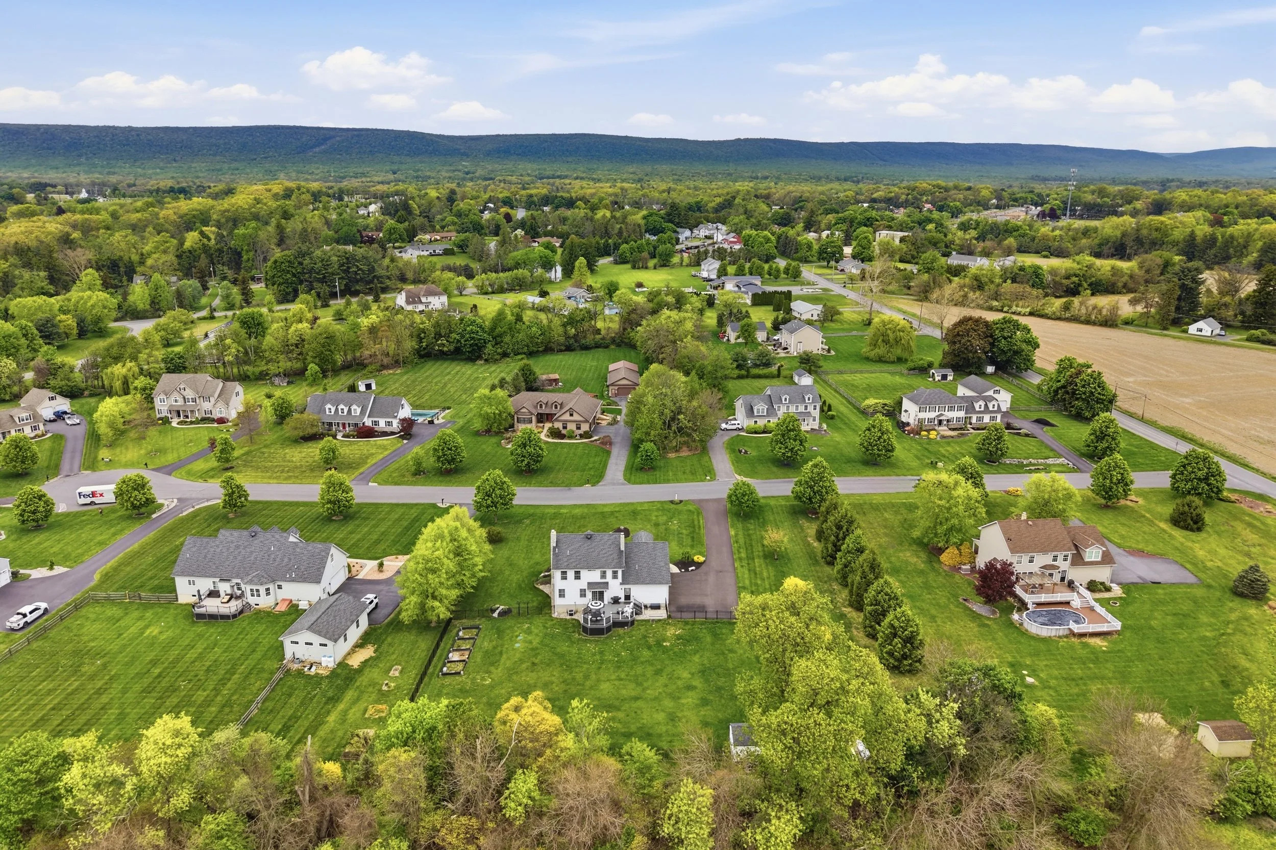 Aerial view of a suburban neighborhood with houses, green lawns, trees, and fields, with a mountain range in the background under a partly cloudy sky.