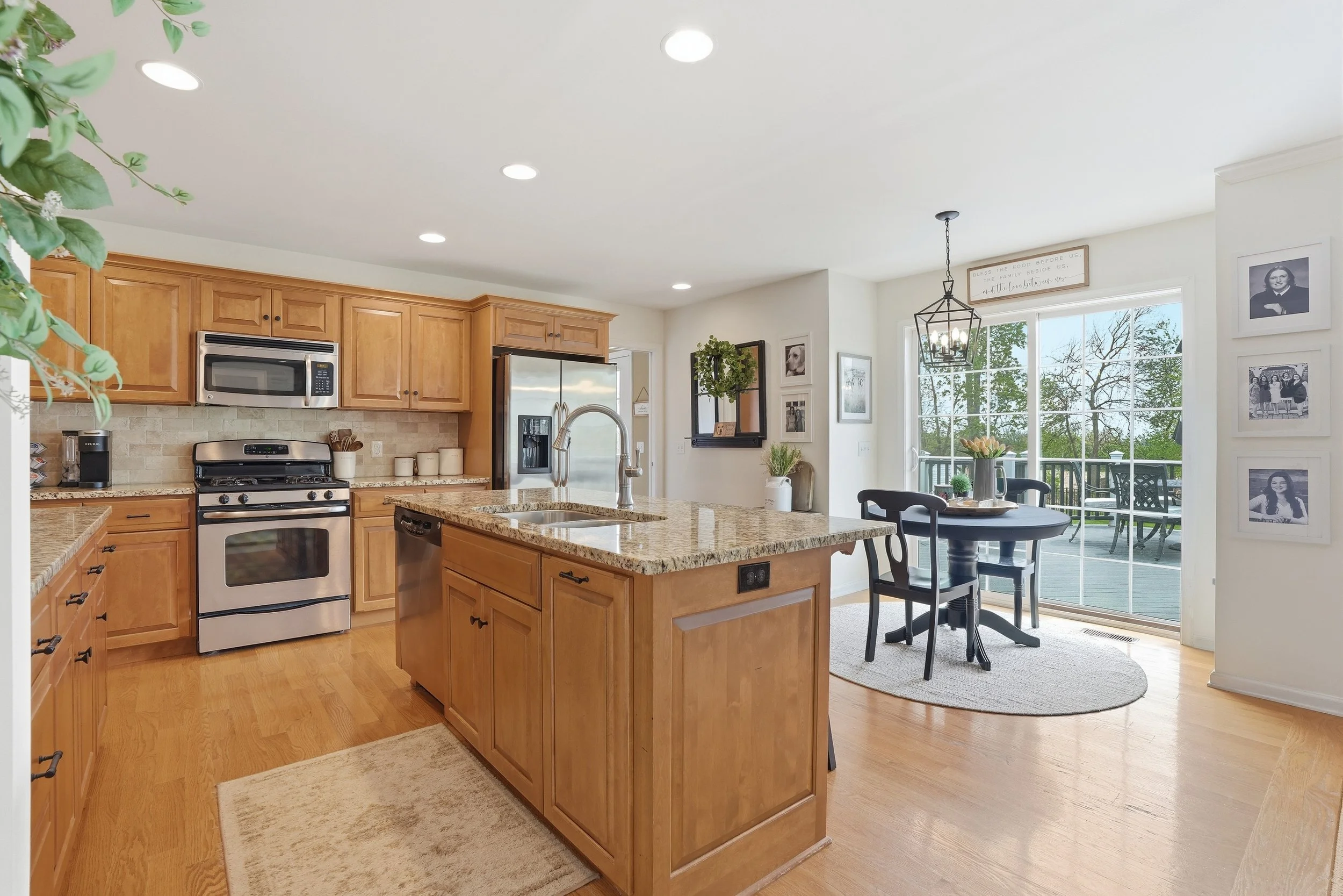 Kitchen with wooden cabinets, stainless steel appliances, granite island, and a dining area by large windows with a view of a deck and greenery outside.