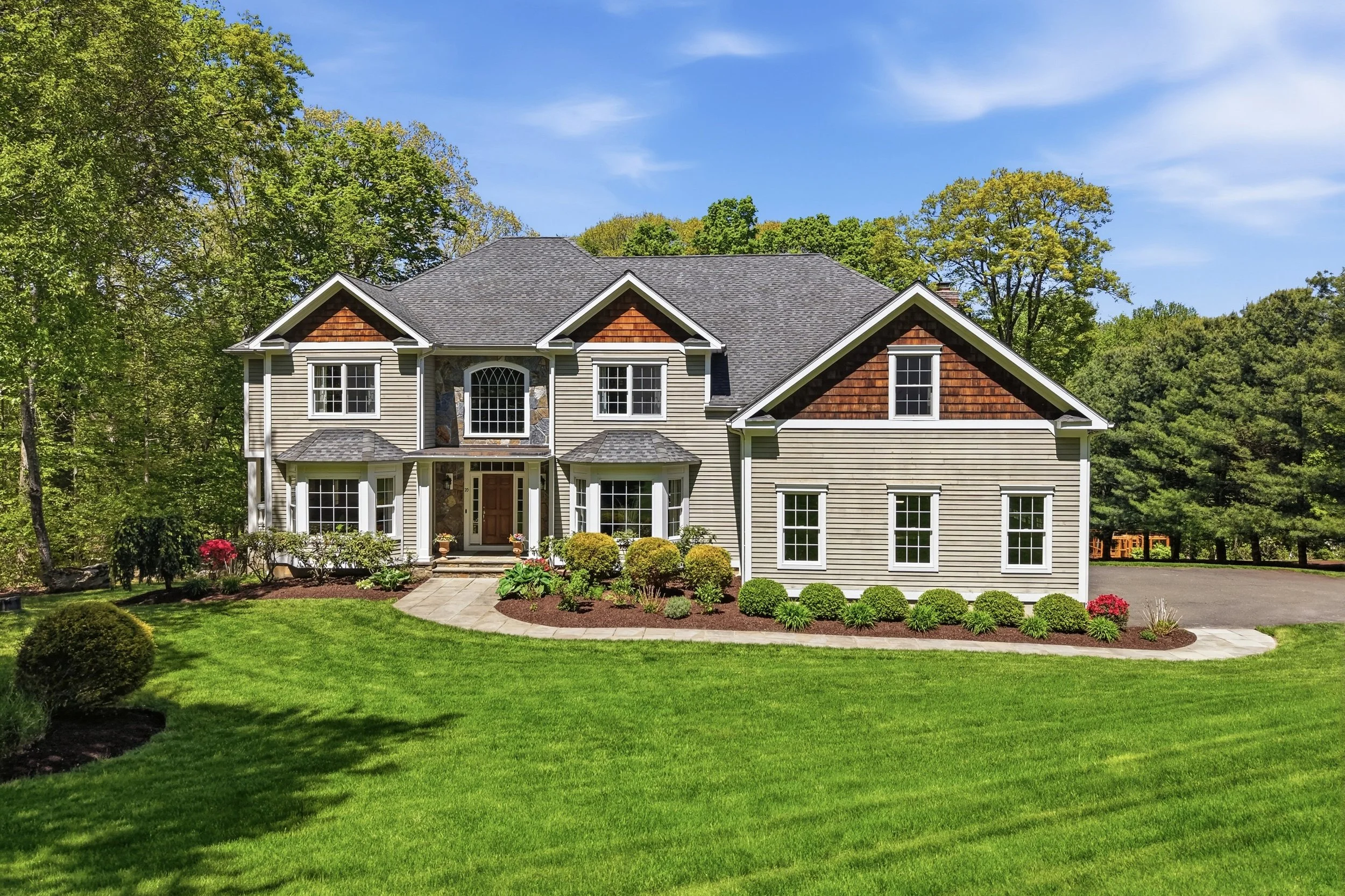 A large two-story house with beige siding, a gable roof, and decorative wooden shingles on the gables. The house has multiple white-framed windows and a front door with a small porch. There is a well-maintained green lawn with bushes, flowers, and a 