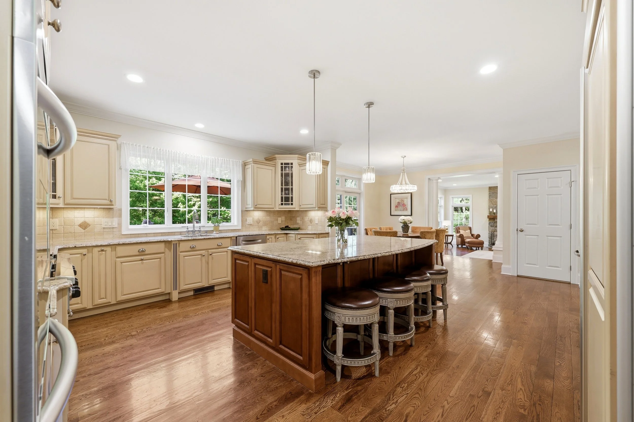 A spacious kitchen with cream-colored cabinets, a granite island with wooden base, and three chairs. There are large windows with curtains, and pendant lights hanging from the ceiling. The background includes a dining area and living room with a chan