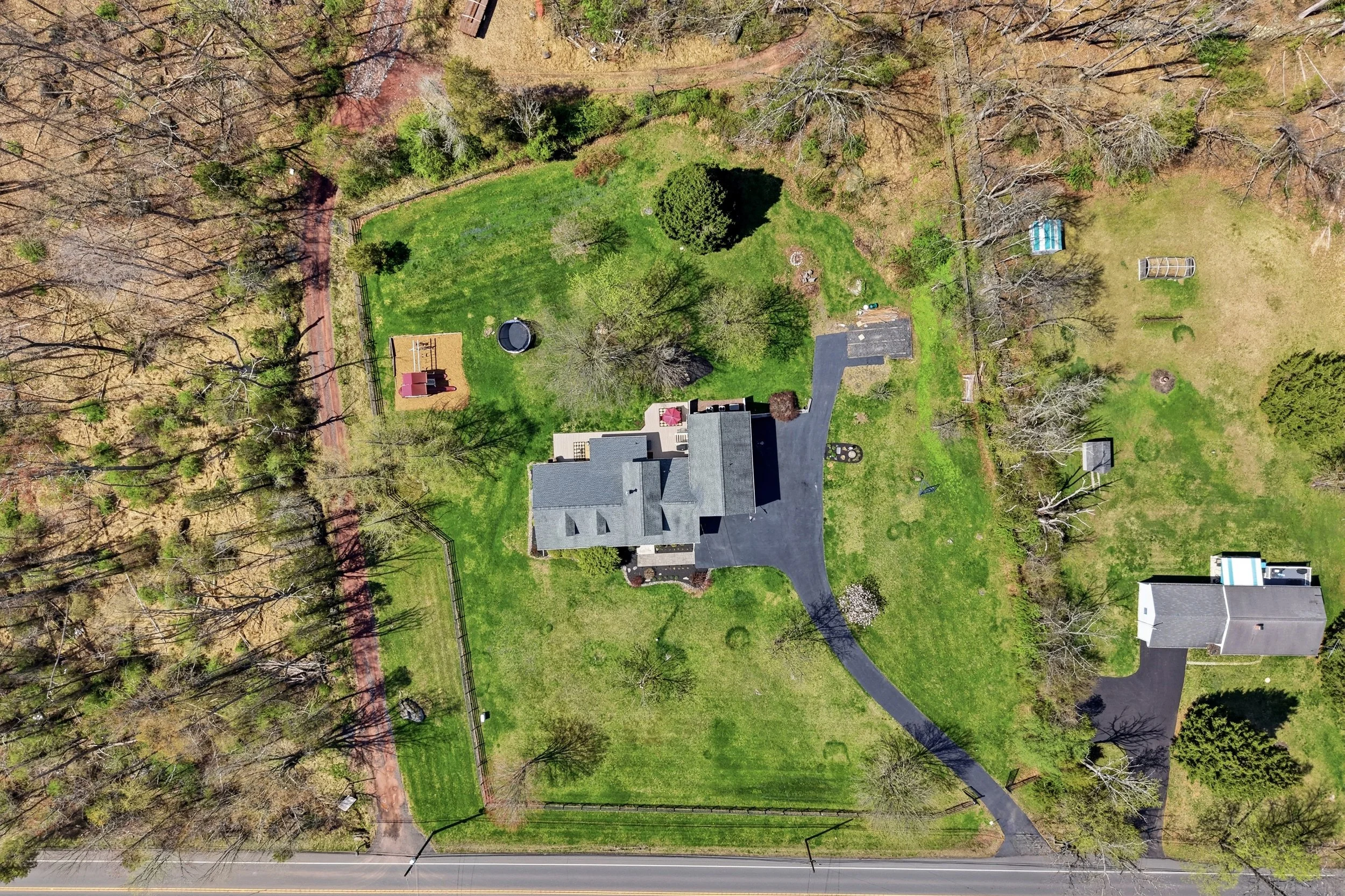An aerial view of a residential backyard with a green lawn, trees, a shed, a trampoline, a slab patio with an umbrella, and a driveway leading to a house, with leafless trees and a road at the bottom.