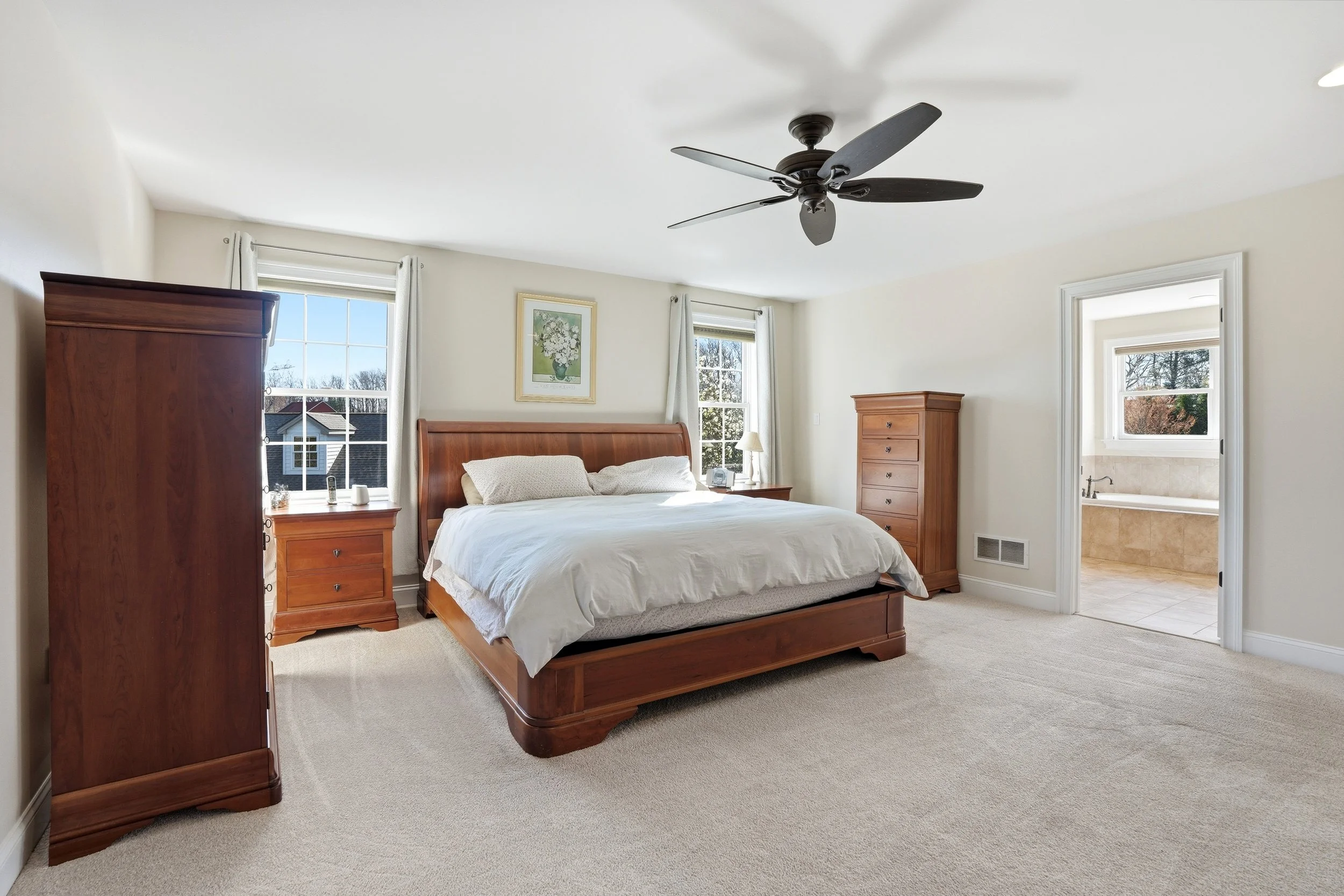 A bedroom with a wooden bed, two matching wooden dressers, and a framed floral painting on the wall. The room has windows with white curtains, and a ceiling fan overhead. Through the doorway, a bathroom is visible.