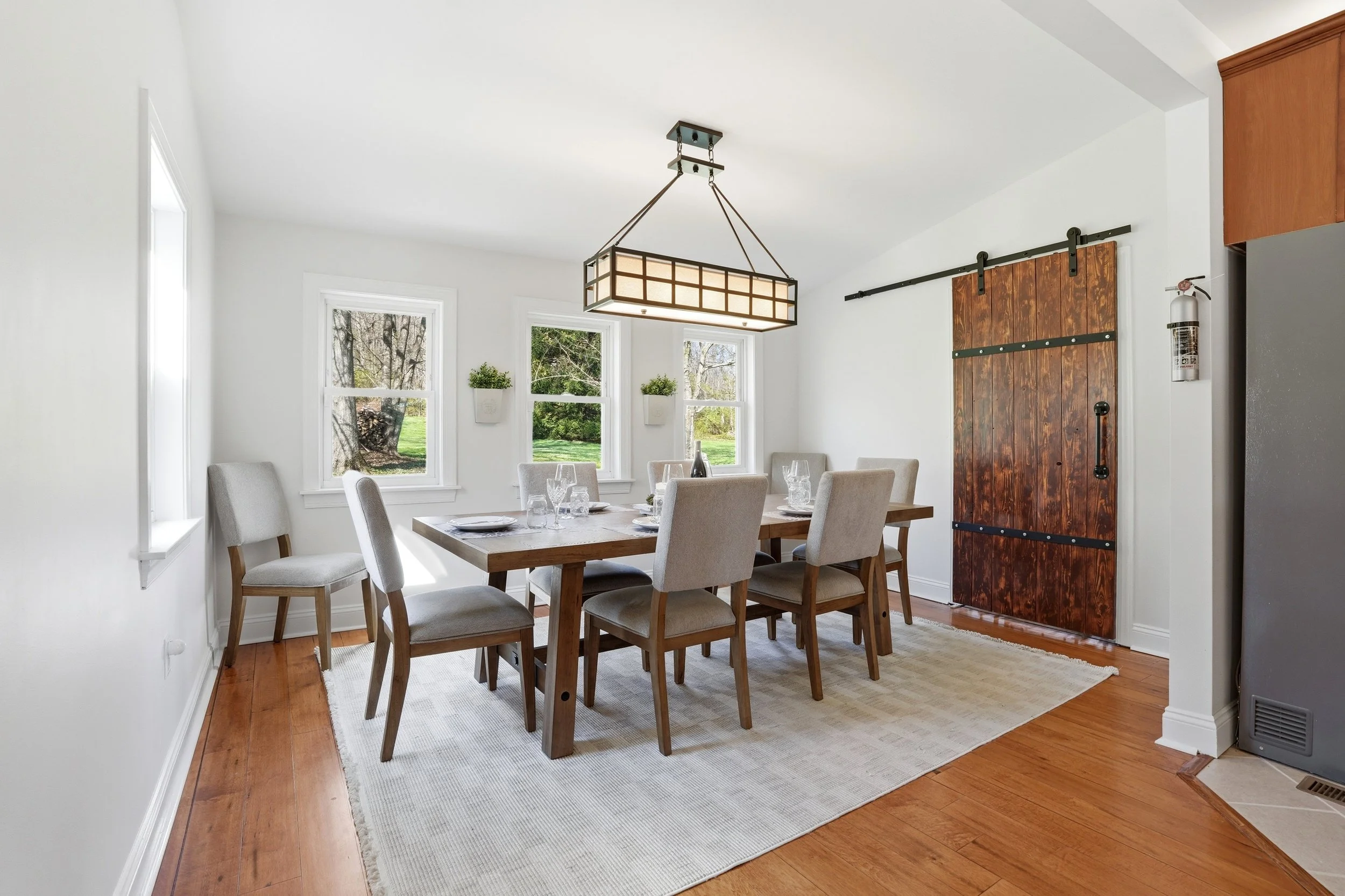 A bright dining room with a wooden table surrounded by six beige upholstered chairs. There are three windows with white trim letting in natural light, and the table is set with plates and wine glasses. A rustic wooden barn door on a sliding track is 