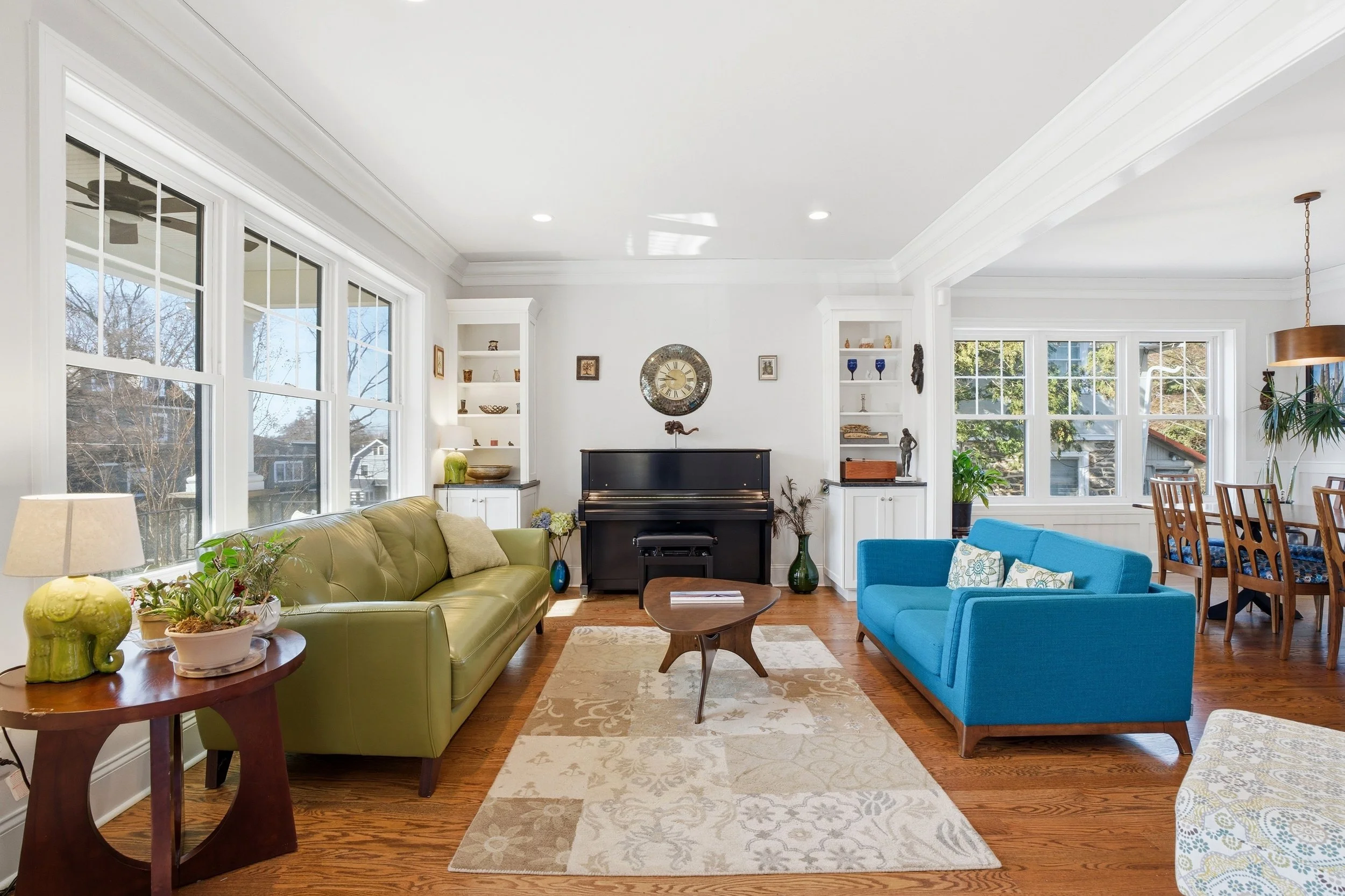 Living room with large windows, white walls, hardwood floor, a black piano, a green sofa with cushions, a blue sofa, small tables with plants, a rug, and decorative items on shelves.