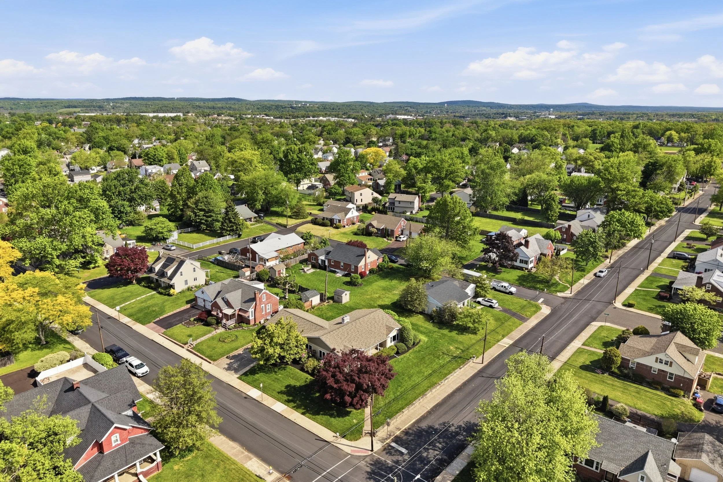 Aerial view of a suburban neighborhood with houses, trees, streets, and distant hills under a partly cloudy sky.