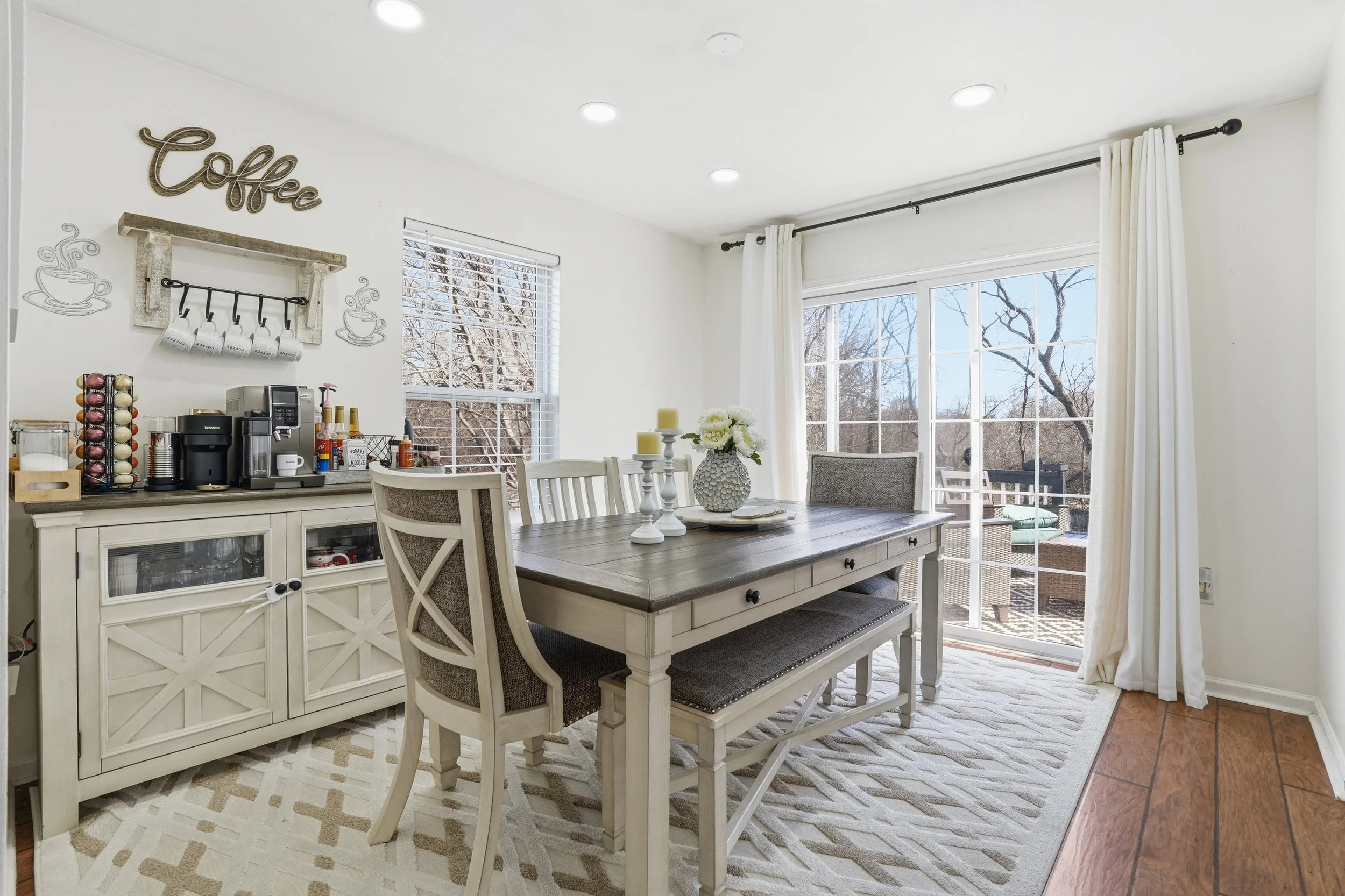 A bright dining room with a wooden table, four chairs, a decorative flower vase, and large windows with white curtains showing trees outside. There's a sideboard with a coffee maker and coffee pod holder, and wall decor including the word 'Coffee' an
