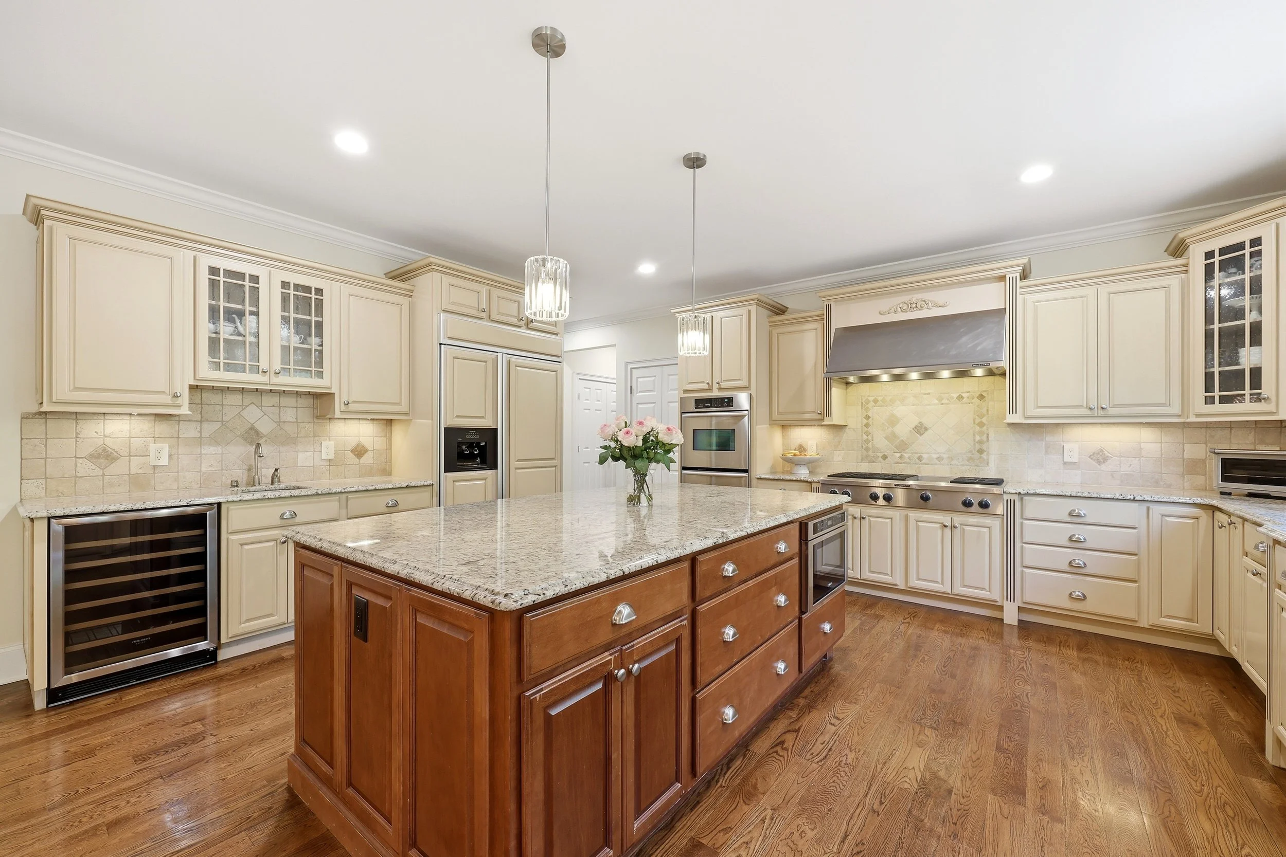 A spacious kitchen with cream-colored cabinets, granite countertops, a central wooden island, built-in microwave, wine cooler, stainless steel oven, and decorative tile backsplash. Two hanging pendant lights and a bouquet of pink flowers are on the i