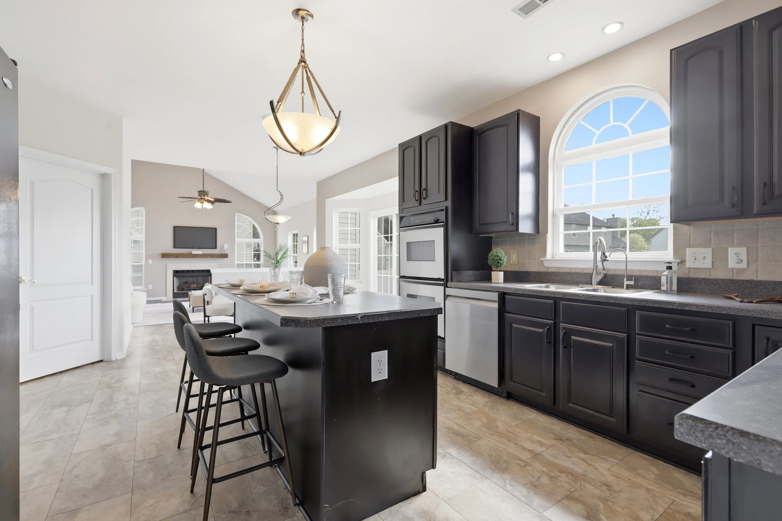 Modern kitchen with black cabinets, granite countertops, a kitchen island with bar stools, large arched window, and a view of a sunny outdoor scene.
