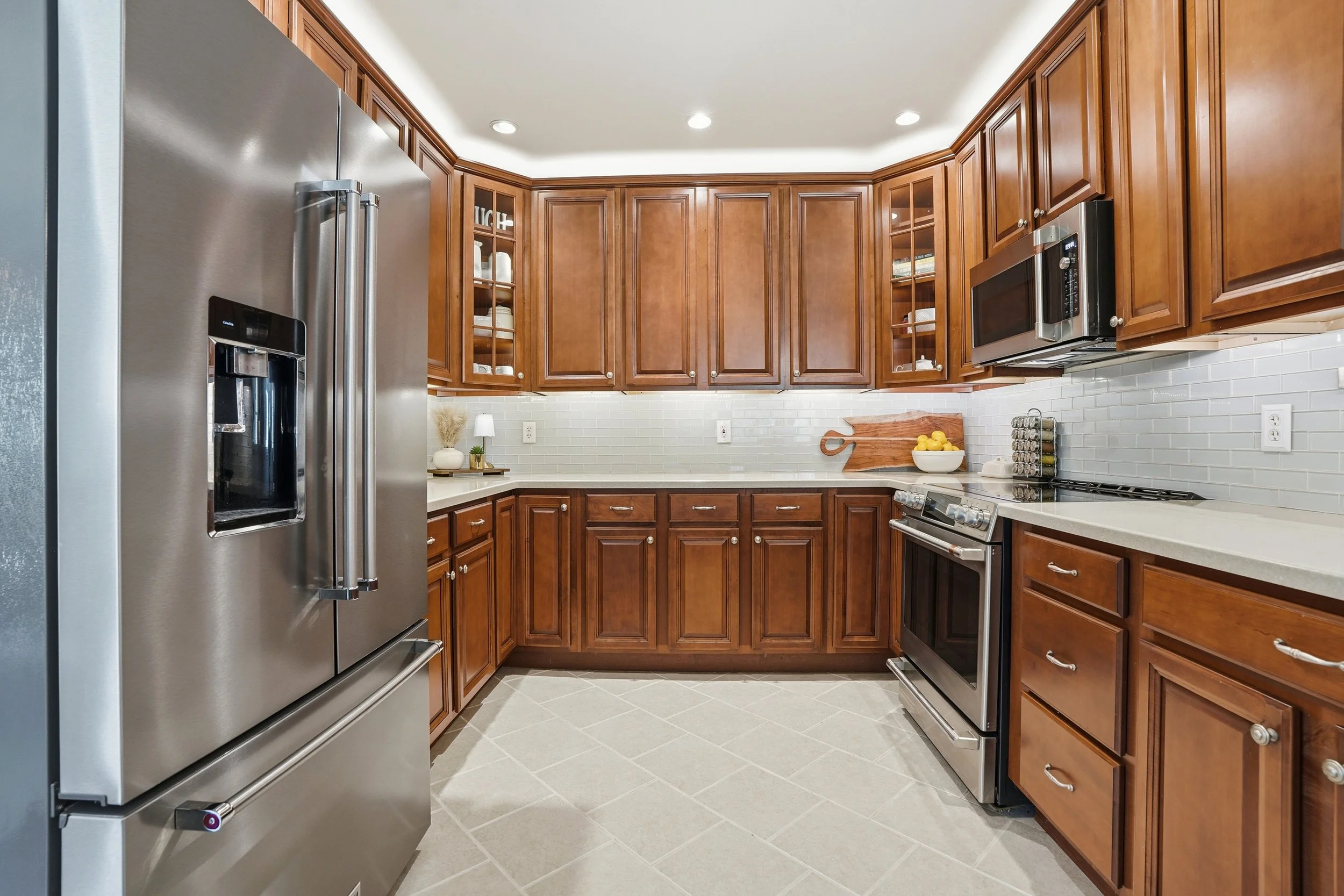 A kitchen with wooden cabinets, white countertops, stainless steel appliances, including a refrigerator, oven, and microwave, on a tile floor.