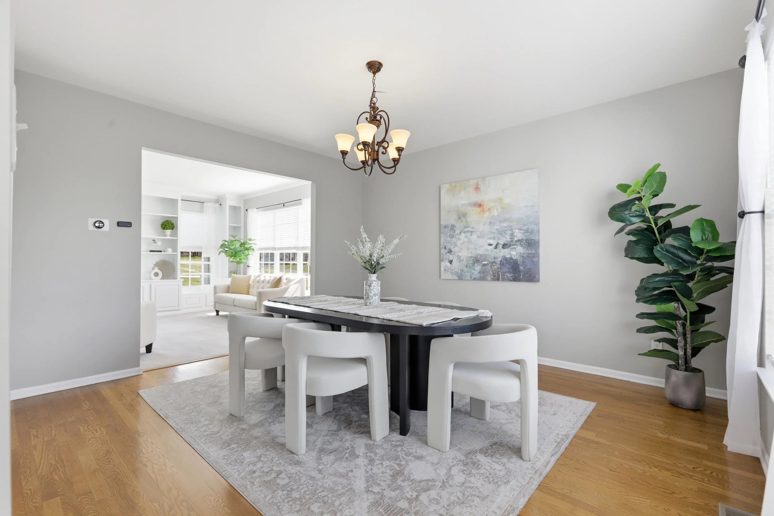 Living and dining area with white walls, a hanging chandelier, a round black dining table with white chairs, a white sofa, art on the wall, a large potted plant, and windows with white curtains.