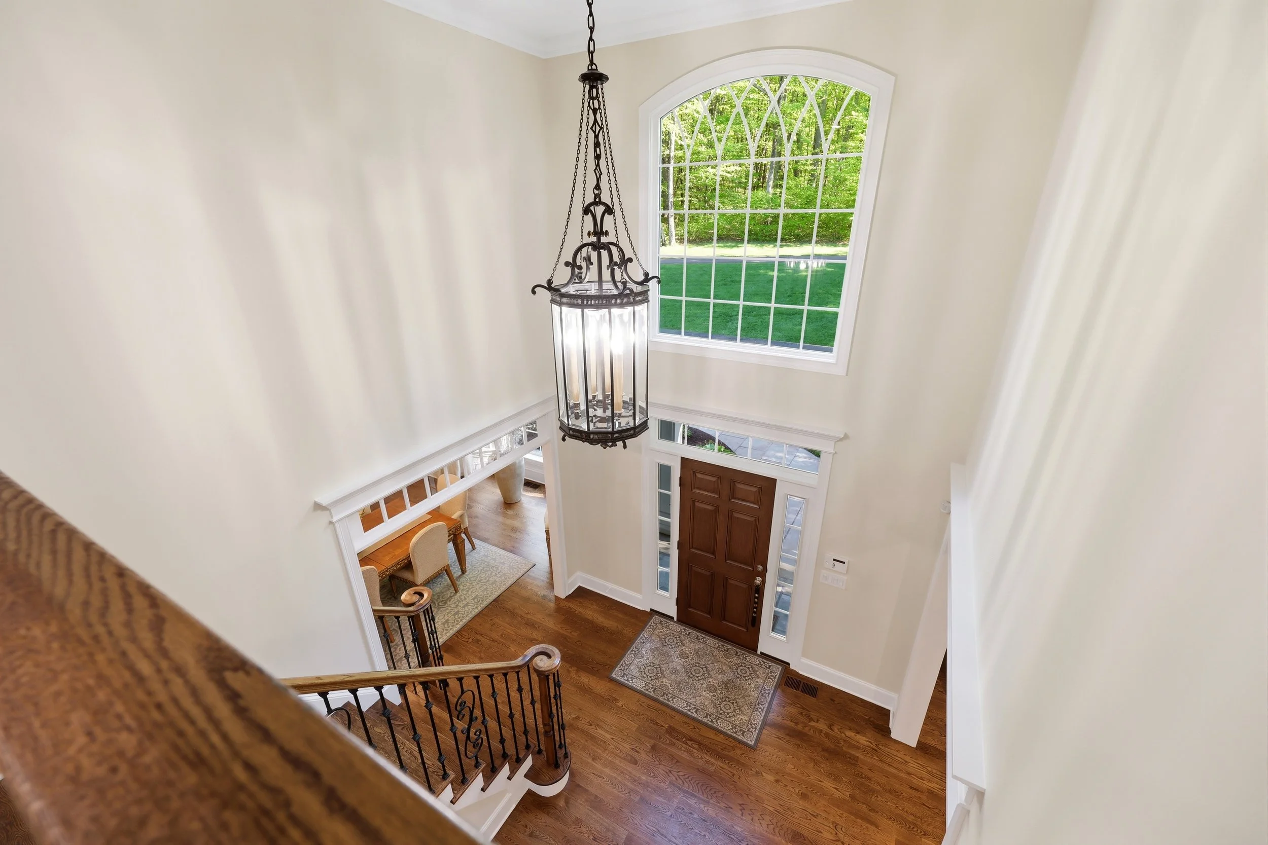 View of a spacious foyer with a wooden front door, large arched window, chandelier, staircase, and a dining area visible in the background.