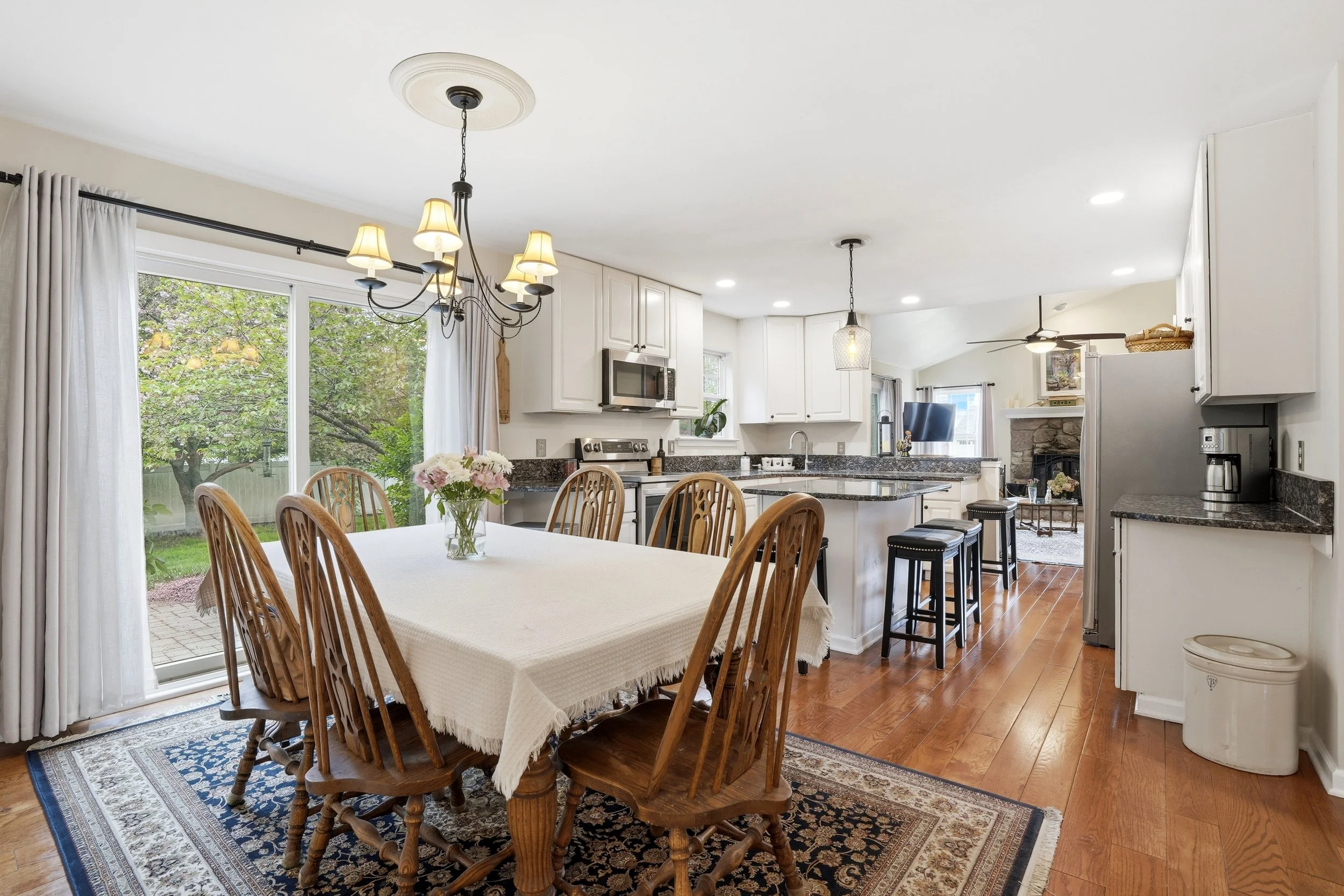 A bright kitchen and dining area with white cabinetry, black granite countertops, a dining table with wooden chairs, and large windows with white curtains showing greenery outside.
