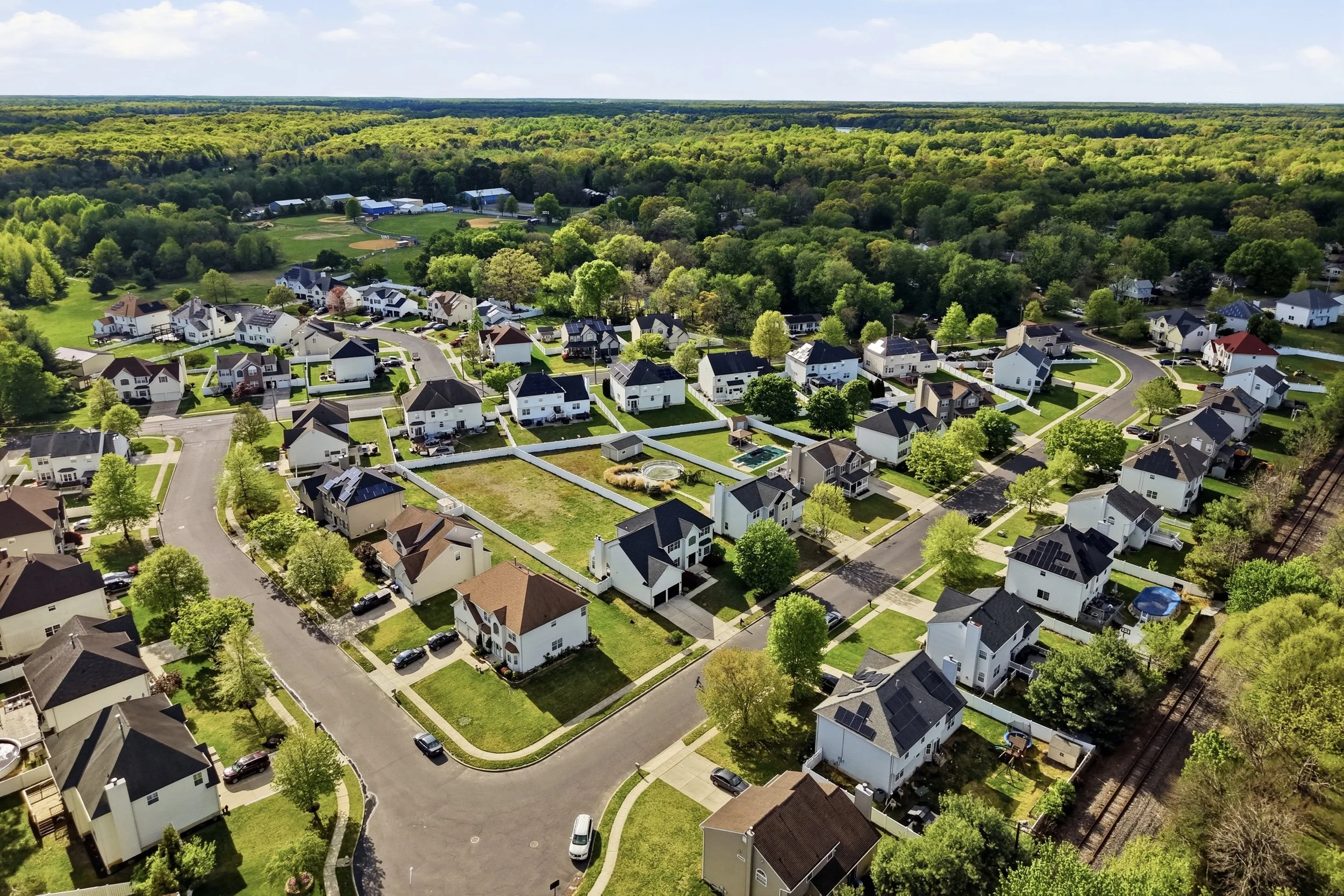 Aerial view of a suburban neighborhood with houses, trees, and streets, adjacent to a wooded area and a baseball field in the distance.