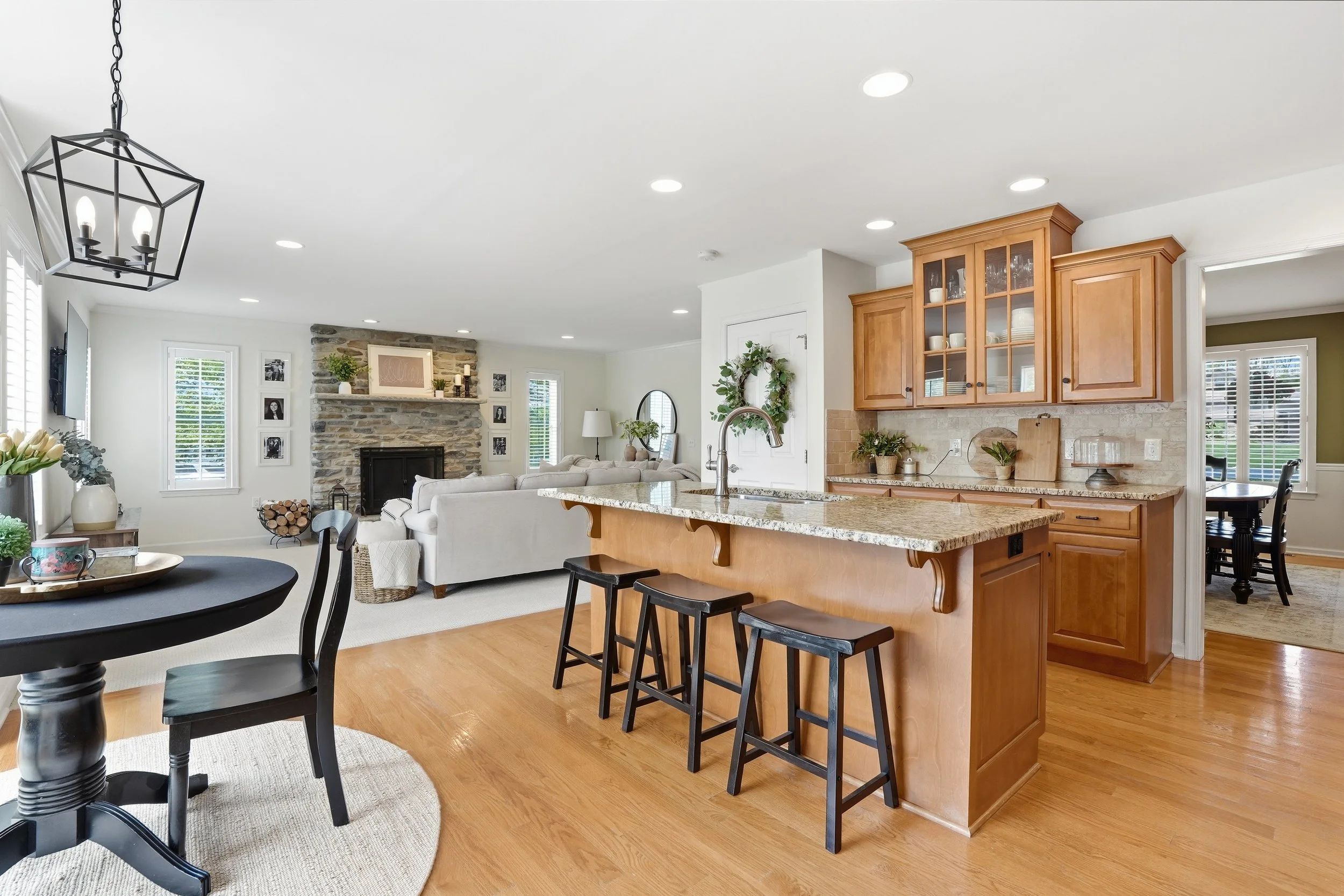 Interior view of a modern open-concept living space featuring a kitchen with wooden cabinets, a granite island with bar stools, a cozy living area with a fireplace, framed photos on the wall, and large windows bringing in natural light.