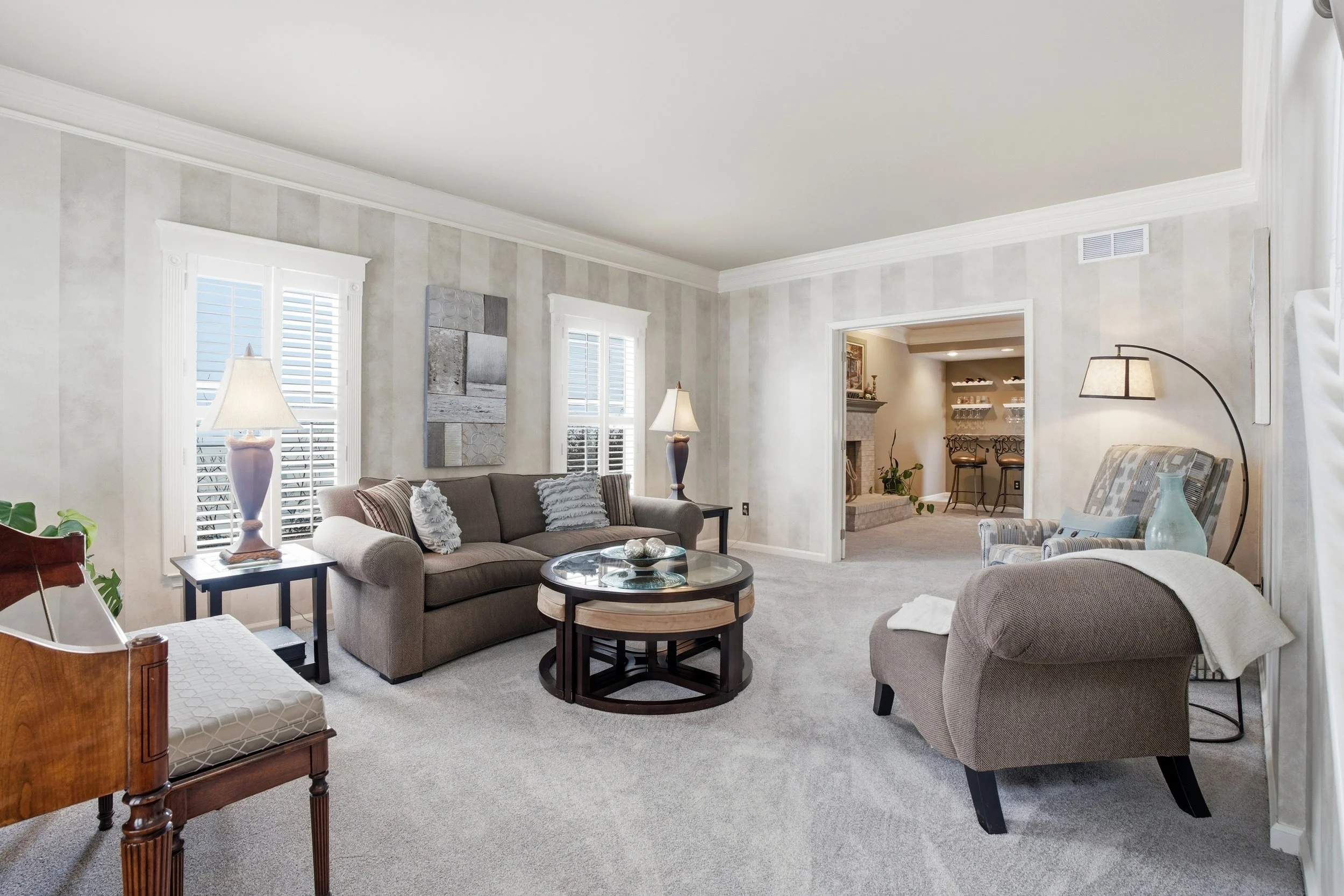 Living room with beige sofas, gray and patterned armchair, coffee table, lamps, and wall art, with large windows and a view into a fireplace room.