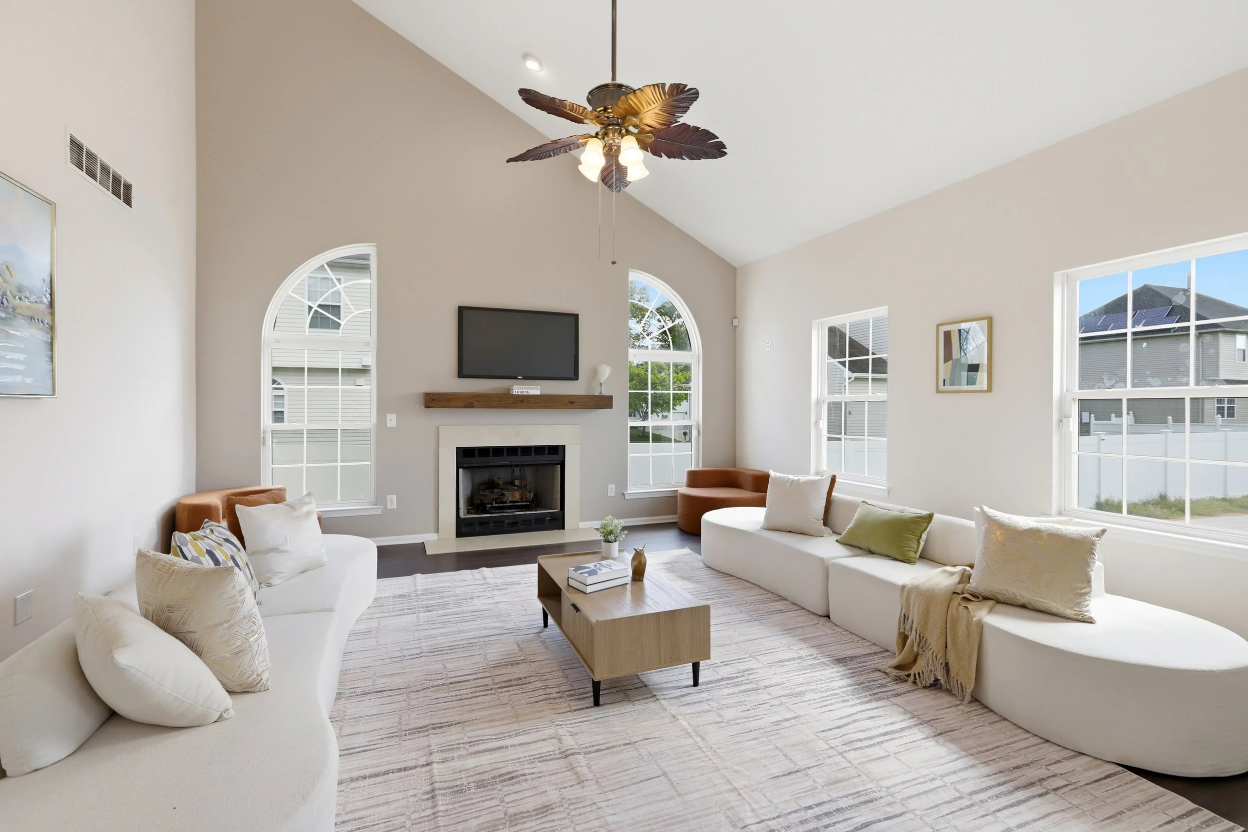 Bright living room with beige walls, multiple windows, a ceiling fan with leaf-shaped blades, a flat-screen TV above a fireplace, and modern furniture including white sofas, a wooden coffee table, and decorative pillows.