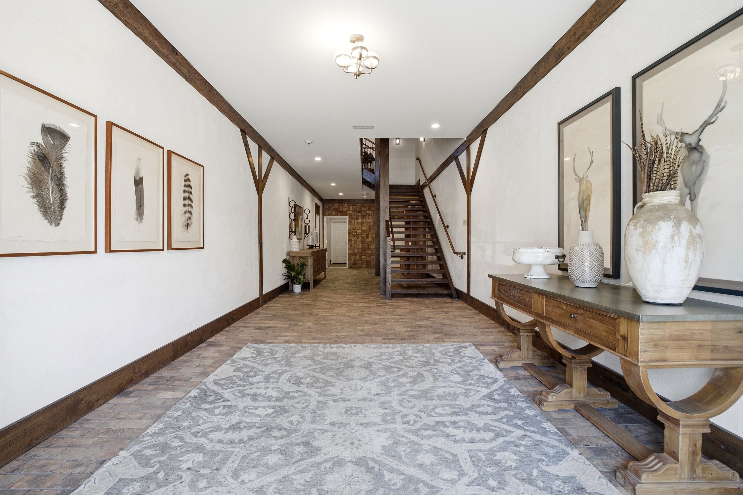A hallway with wooden accents and decor, including framed feathers on the left wall and pottery with dried plants on the right table. There's a staircase at the end with wooden steps and a brick accent wall behind it. The floor is made of brick, and 