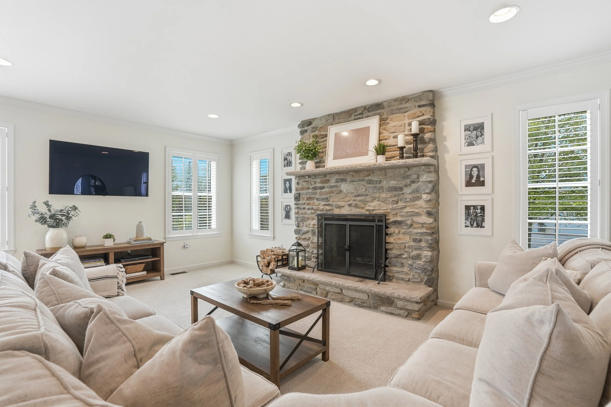 A living room with a stone fireplace, beige sofas, a wooden coffee table, and large windows with white shutters.