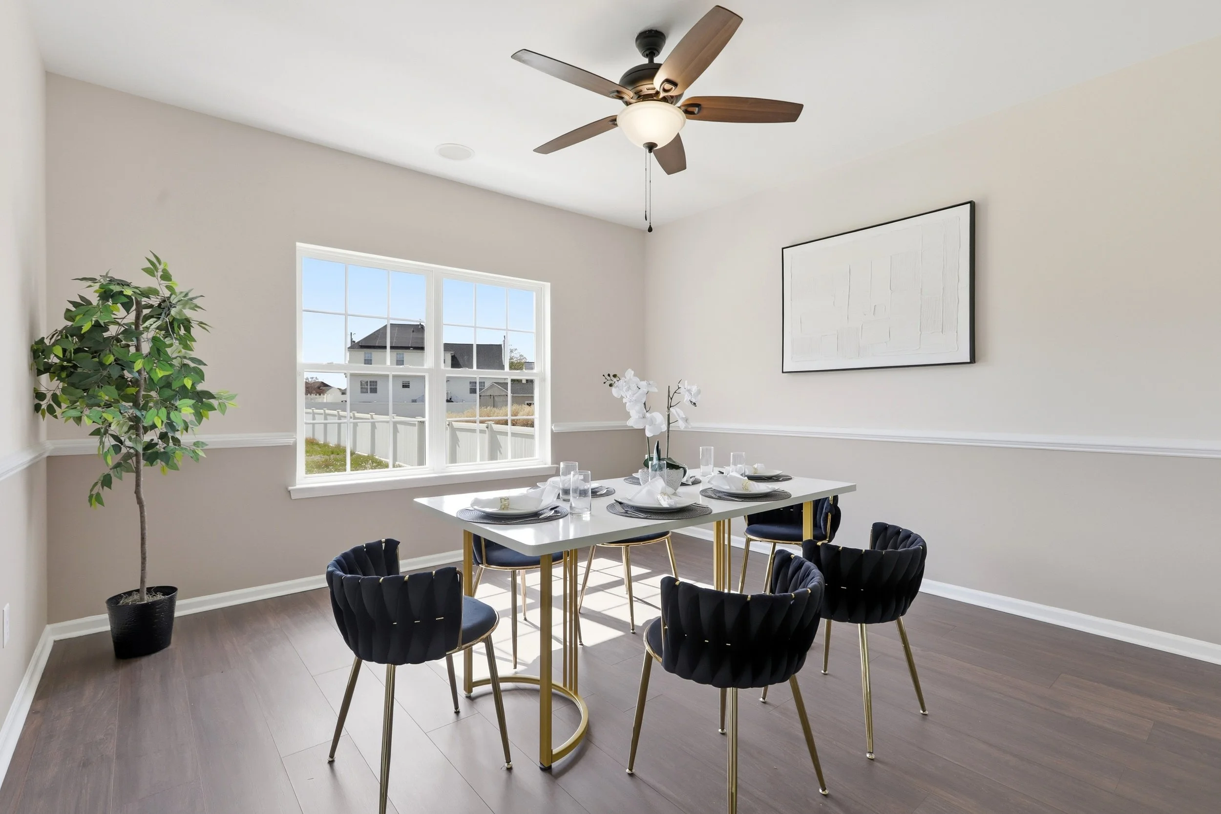A dining room with a white table set with plates, glasses, and napkins, surrounded by six black velvet chairs with gold legs, a potted plant in the corner, large window letting in natural light, and a ceiling fan with wooden blades.