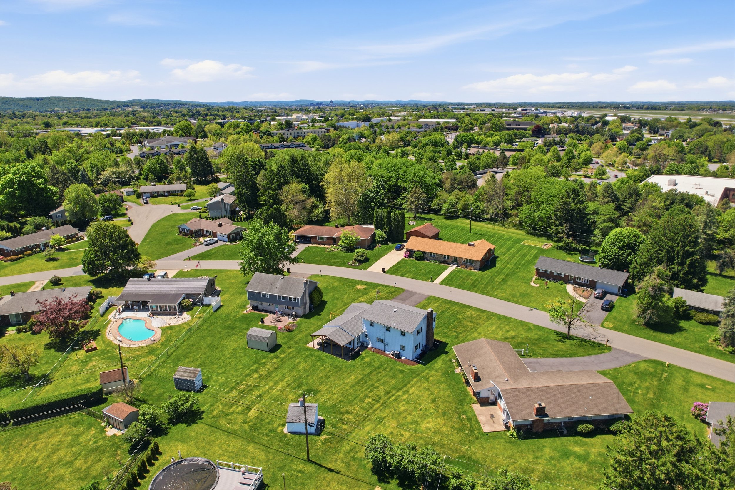 Aerial view of a suburban neighborhood with houses, green lawns, trees, and a swimming pool, under a partly cloudy blue sky.