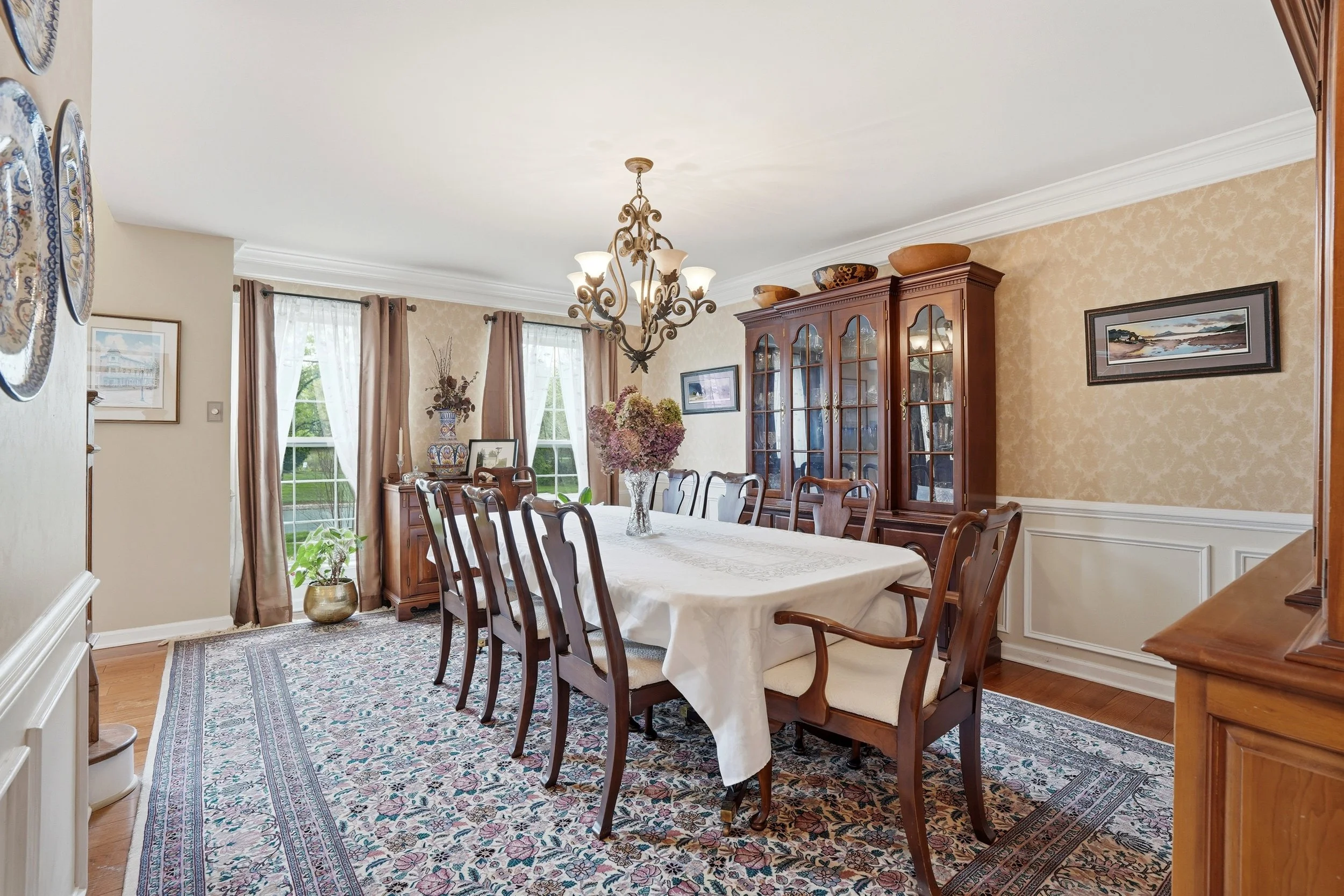 A dining room with a large table covered with a white tablecloth, surrounded by wooden chairs. There is a chandelier hanging above the table, and a china cabinet against the wall. The room has windows with sheer curtains, and decorative items like va