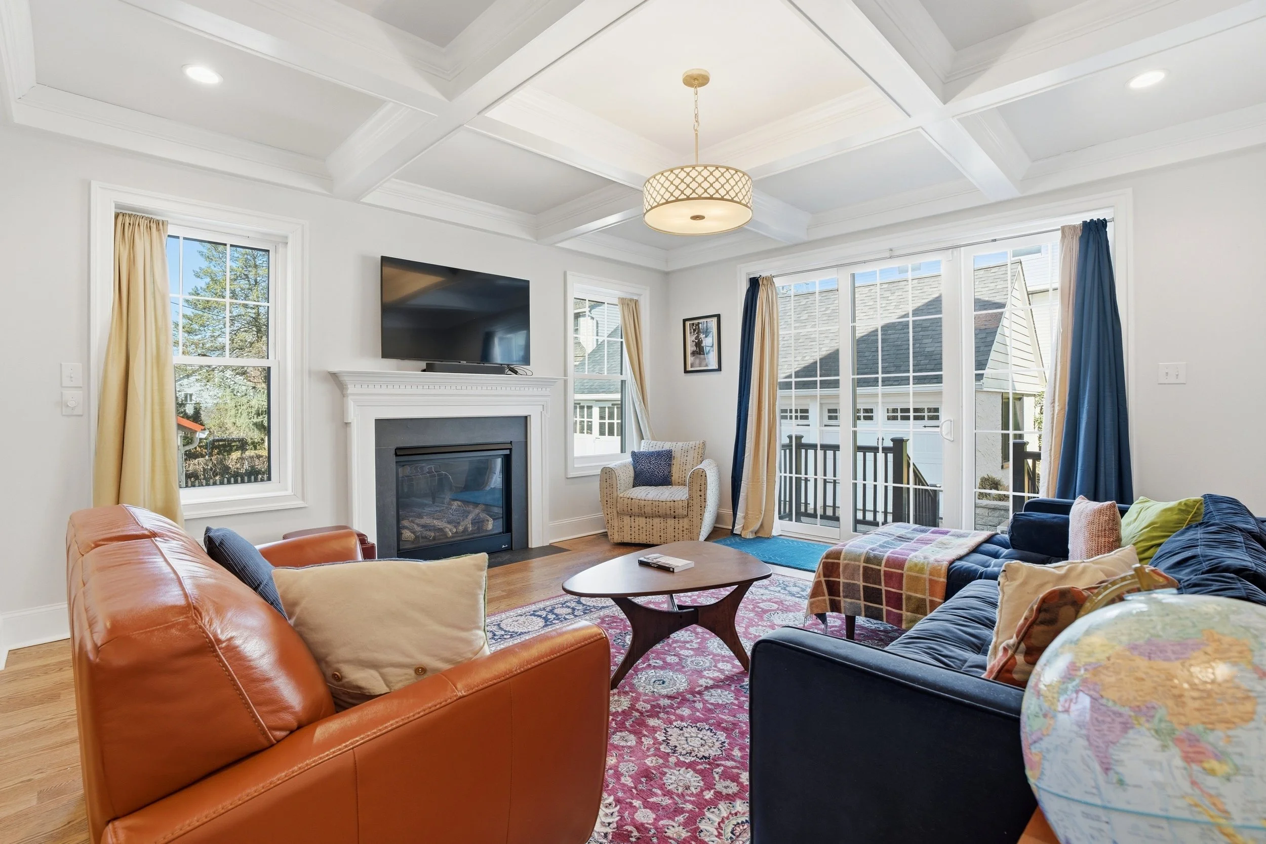 Living room with large windows, a sofa, an armchair, a fireplace with a TV mounted above, and a round table with a world map on it, decorated with colorful curtains and a patterned area rug.
