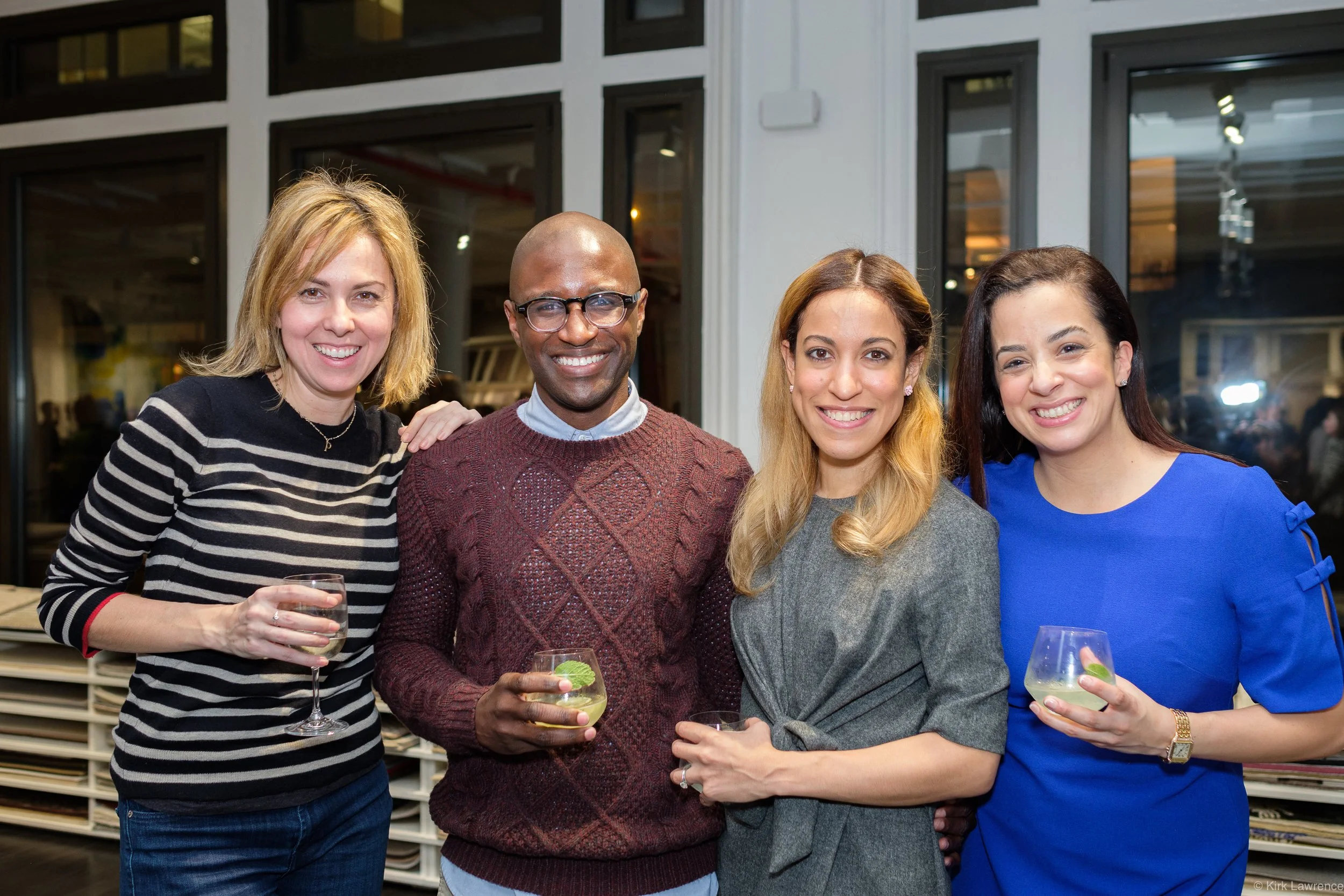 Four women and one man standing together at a social event, holding drinks, smiling for the camera in an indoor setting with large windows in the background.