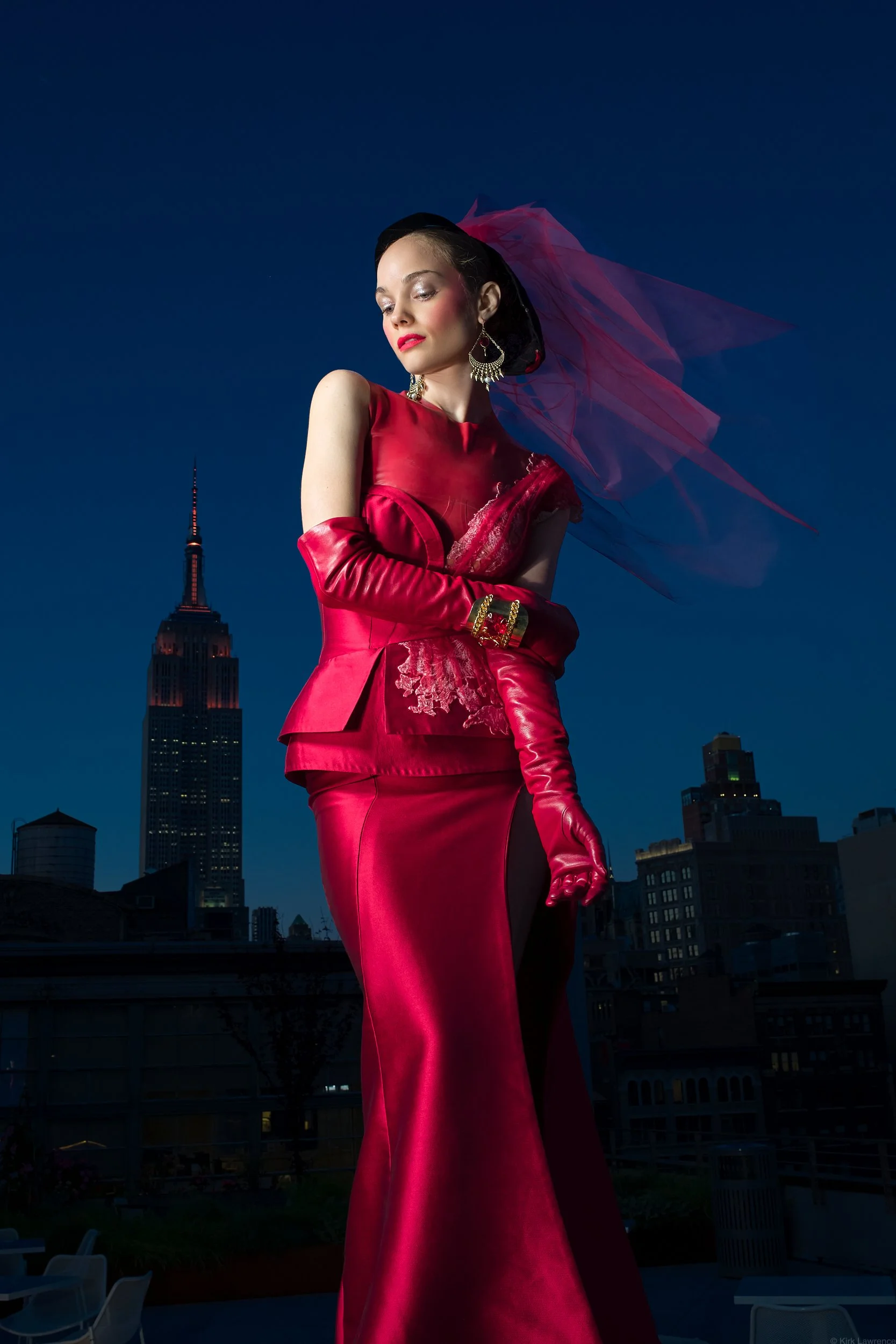 A woman in a red satin evening gown, long red gloves, and a pink veil stands against a nighttime city skyline with the Empire State Building in the background.