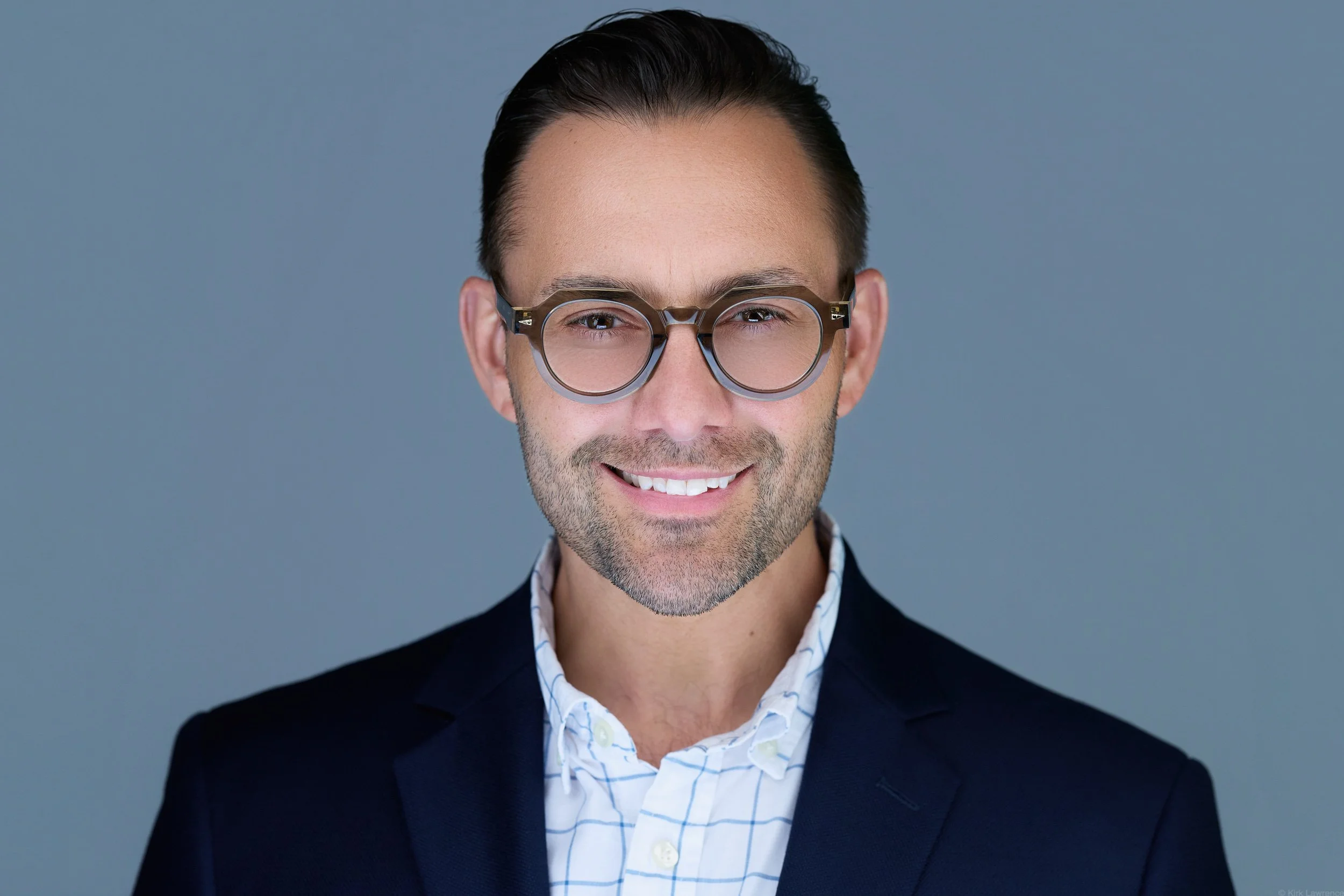 A portrait of a smiling man with dark hair, wearing glasses, a checked shirt, and a dark blazer against a plain grey background.