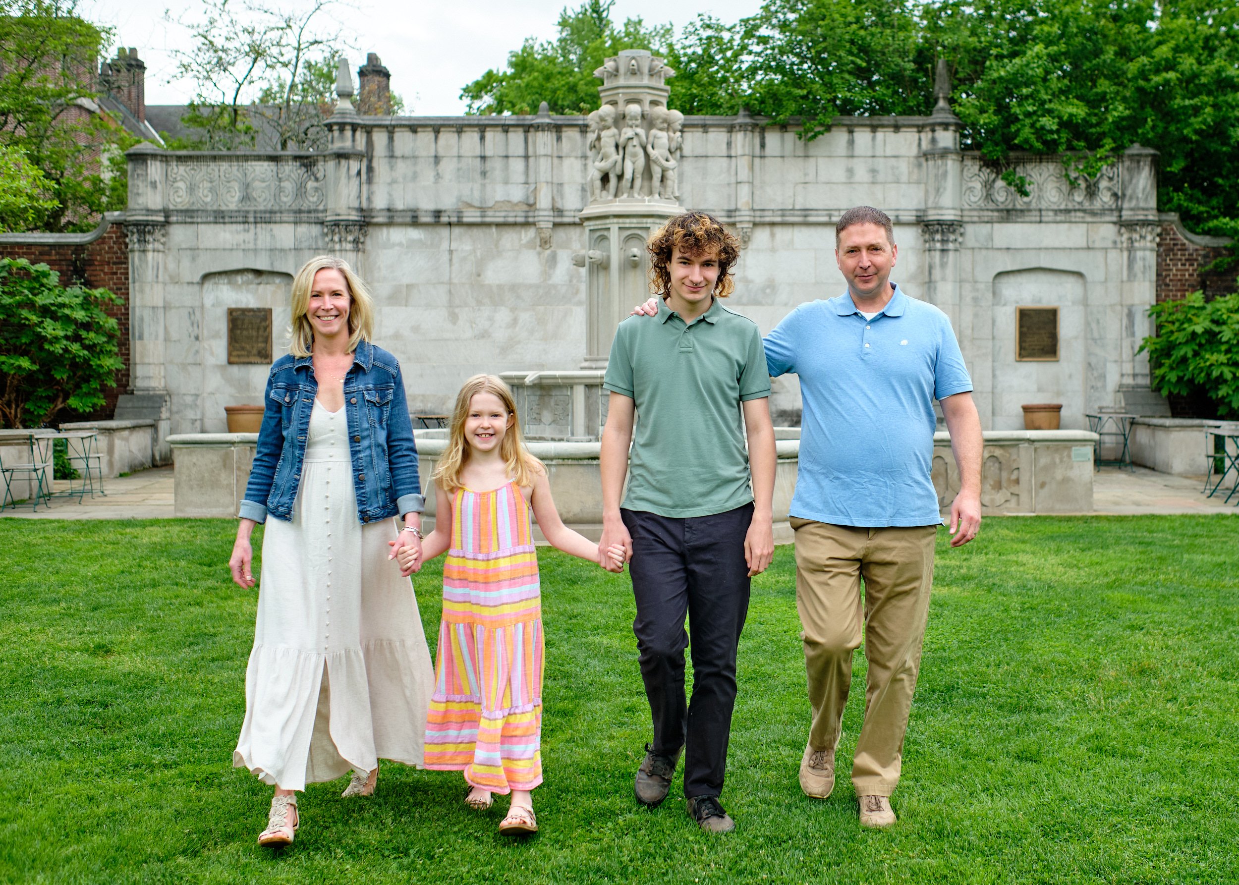 A family of five walking on a green lawn in front of a stone fountain with sculptures, surrounded by trees and historic buildings.