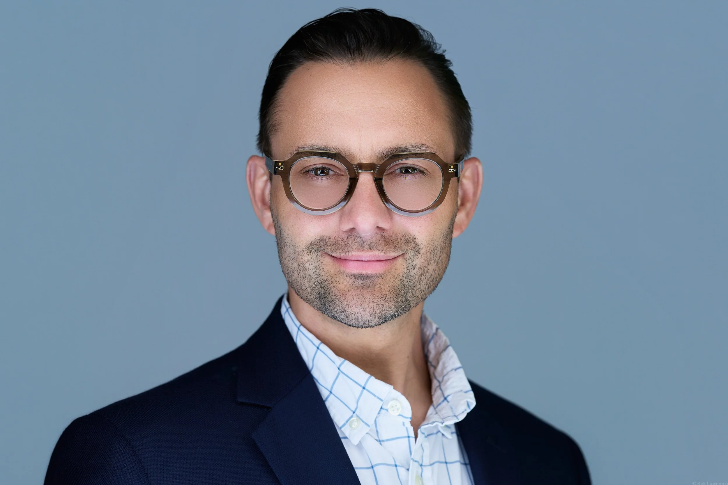Portrait of a man with dark hair, wearing glasses, a white shirt with blue checks, and a dark blazer, against a plain light grey background.