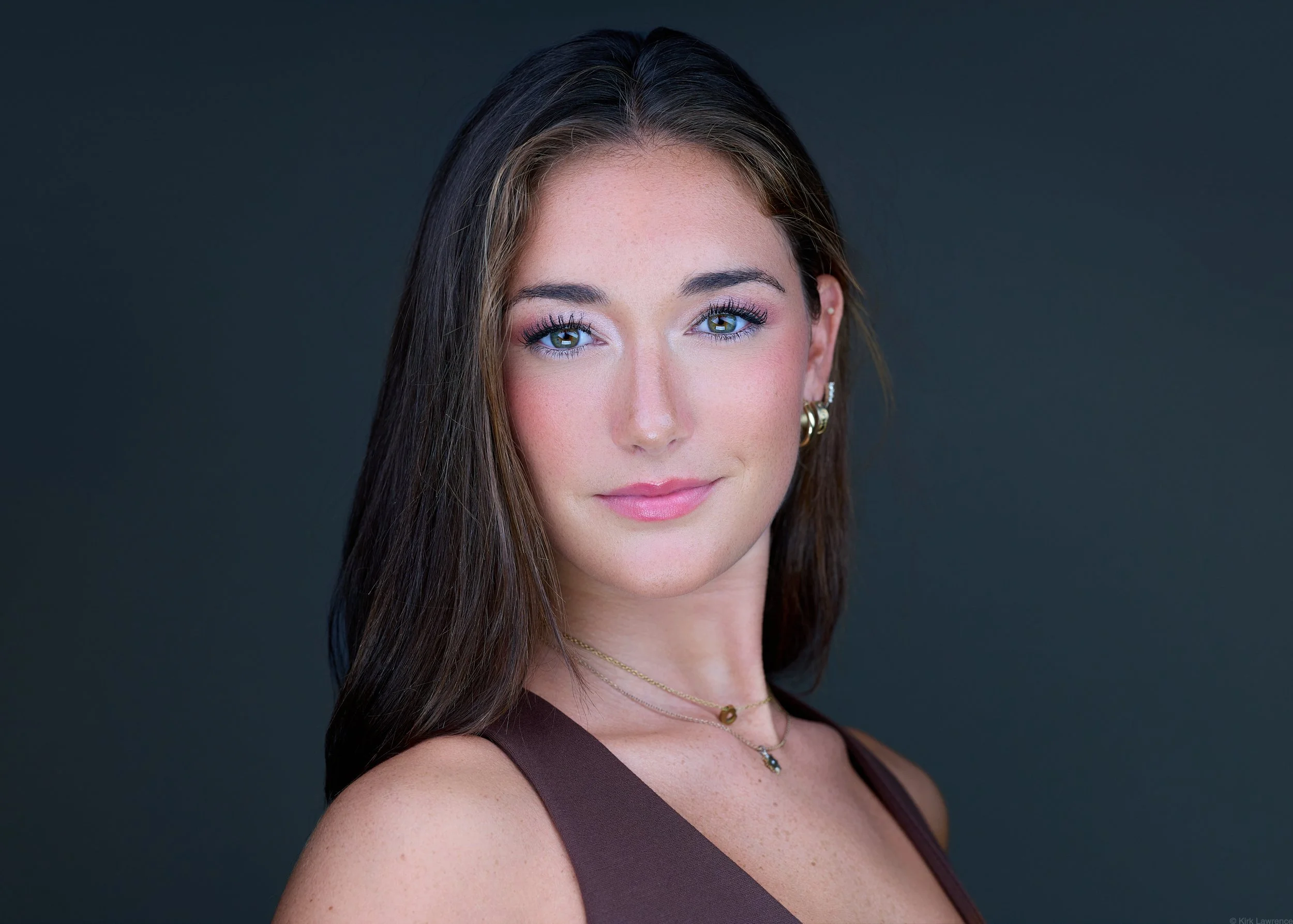 Close-up portrait of a woman with long brown hair, blue eyes, pink makeup, and gold earrings, against a dark gray background.