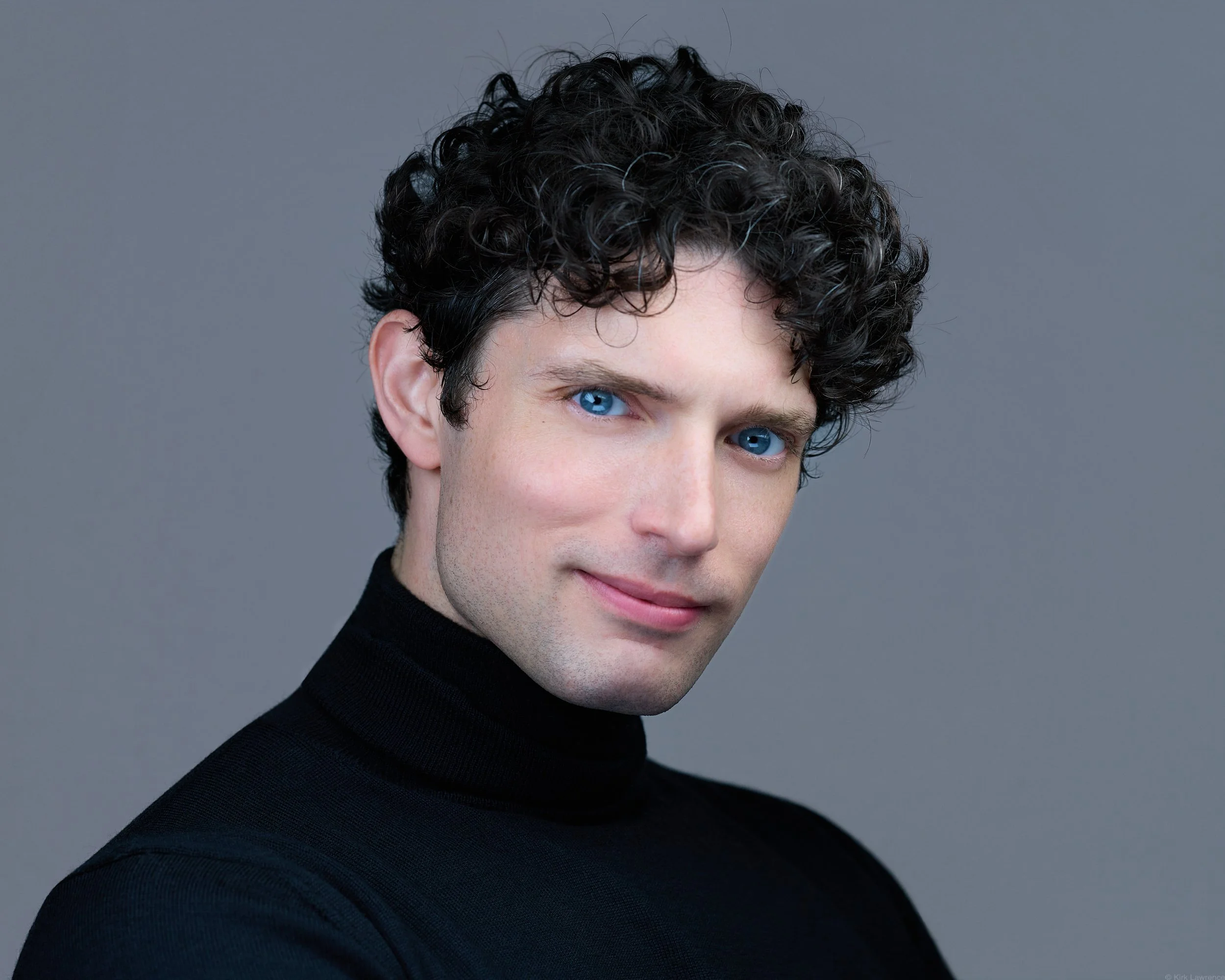 A young man with curly dark hair and bright blue eyes, wearing a black turtleneck sweater, against a plain gray background.