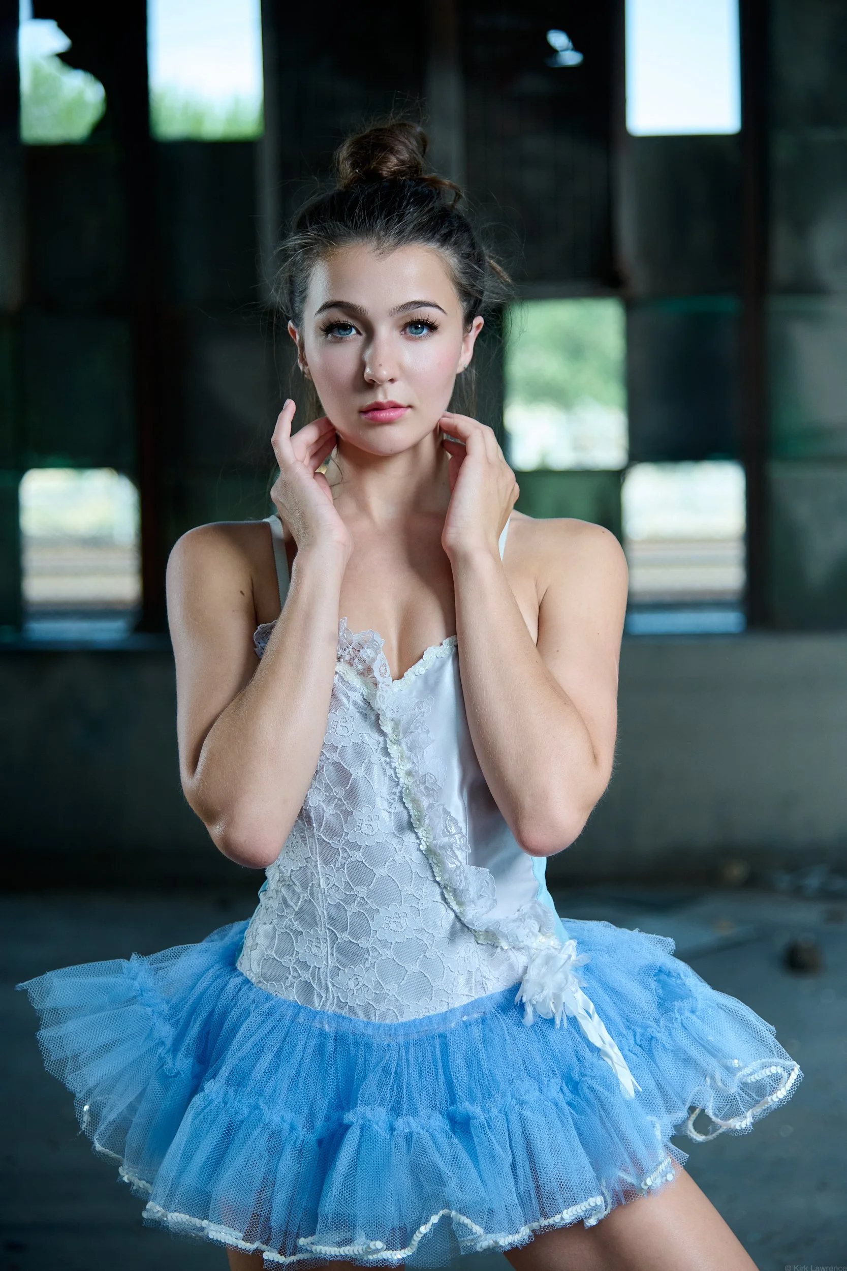 A young woman in a white lace top and blue tutu posing indoors with her hands near her face.
