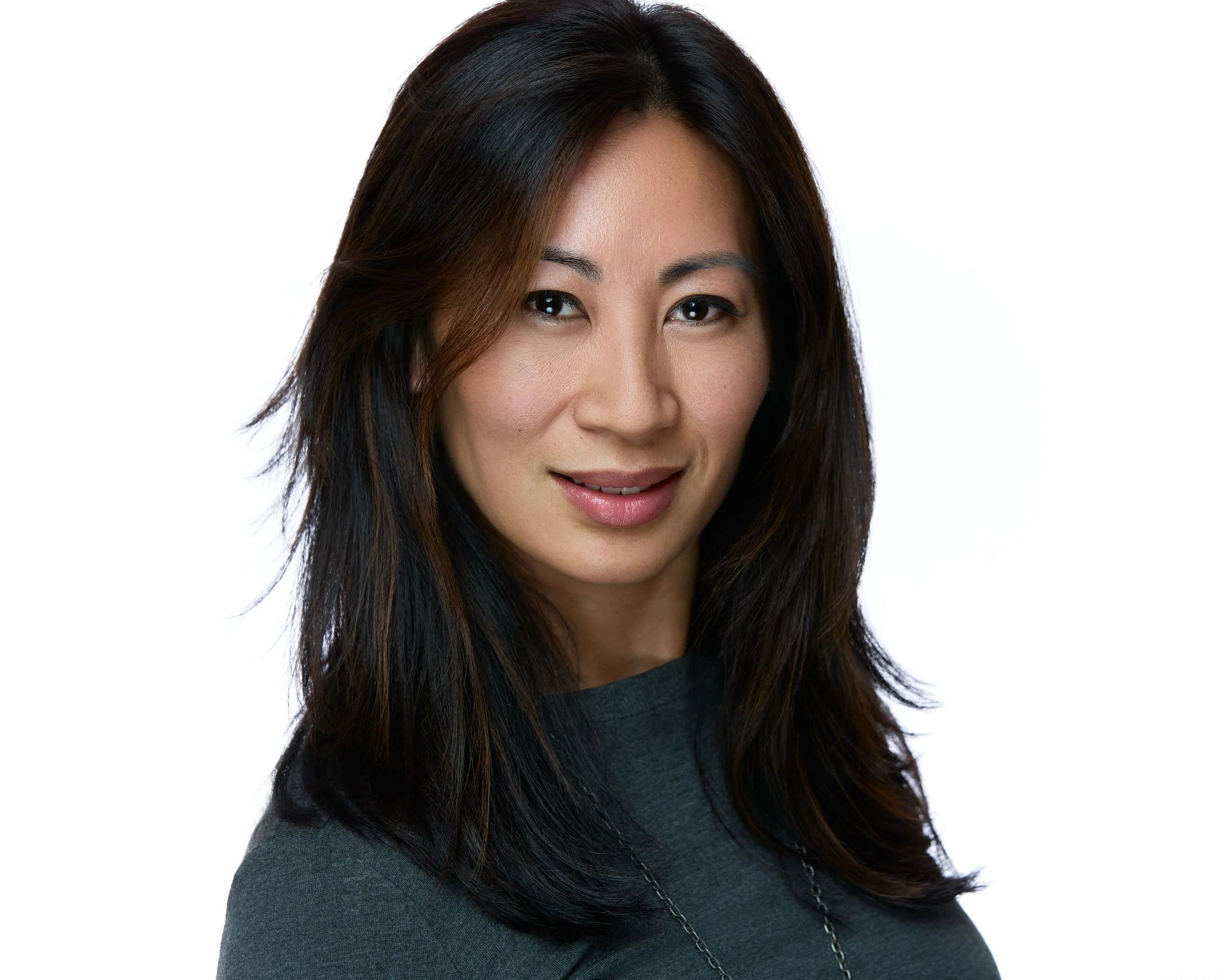 A woman with dark brown hair and subtle makeup, wearing a gray top and a necklace, smiling softly at the camera against a white background.