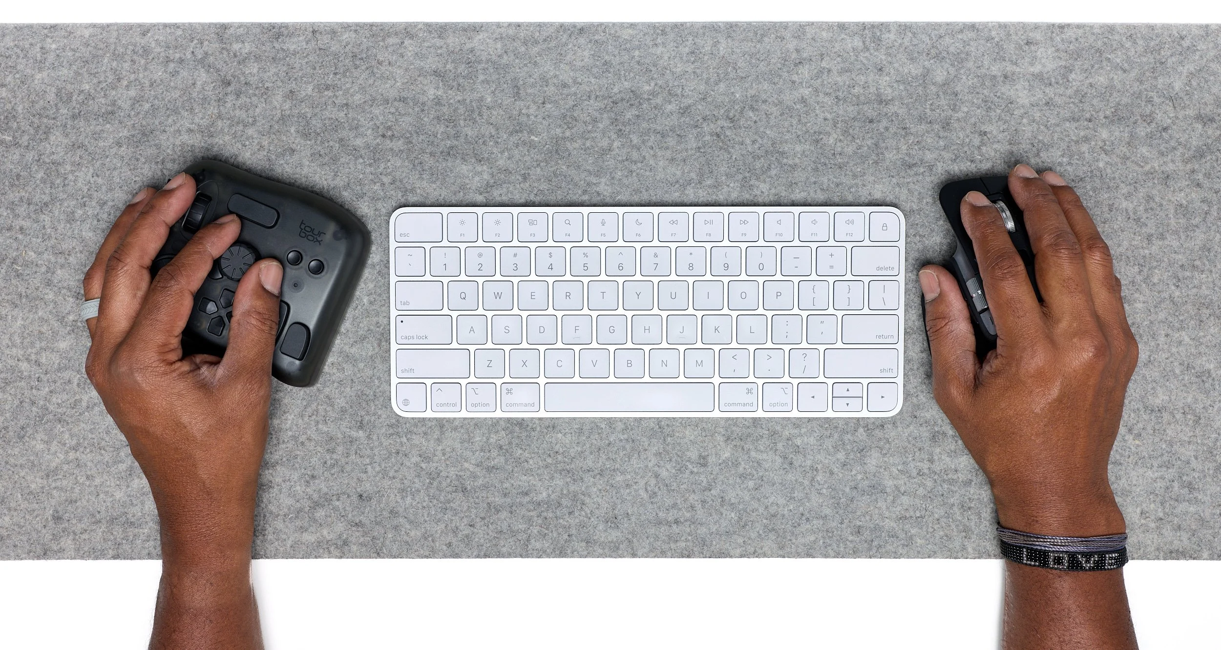 Overhead view of a person's hands operating a computer mouse and a remote control on a gray desk with a white keyboard.
