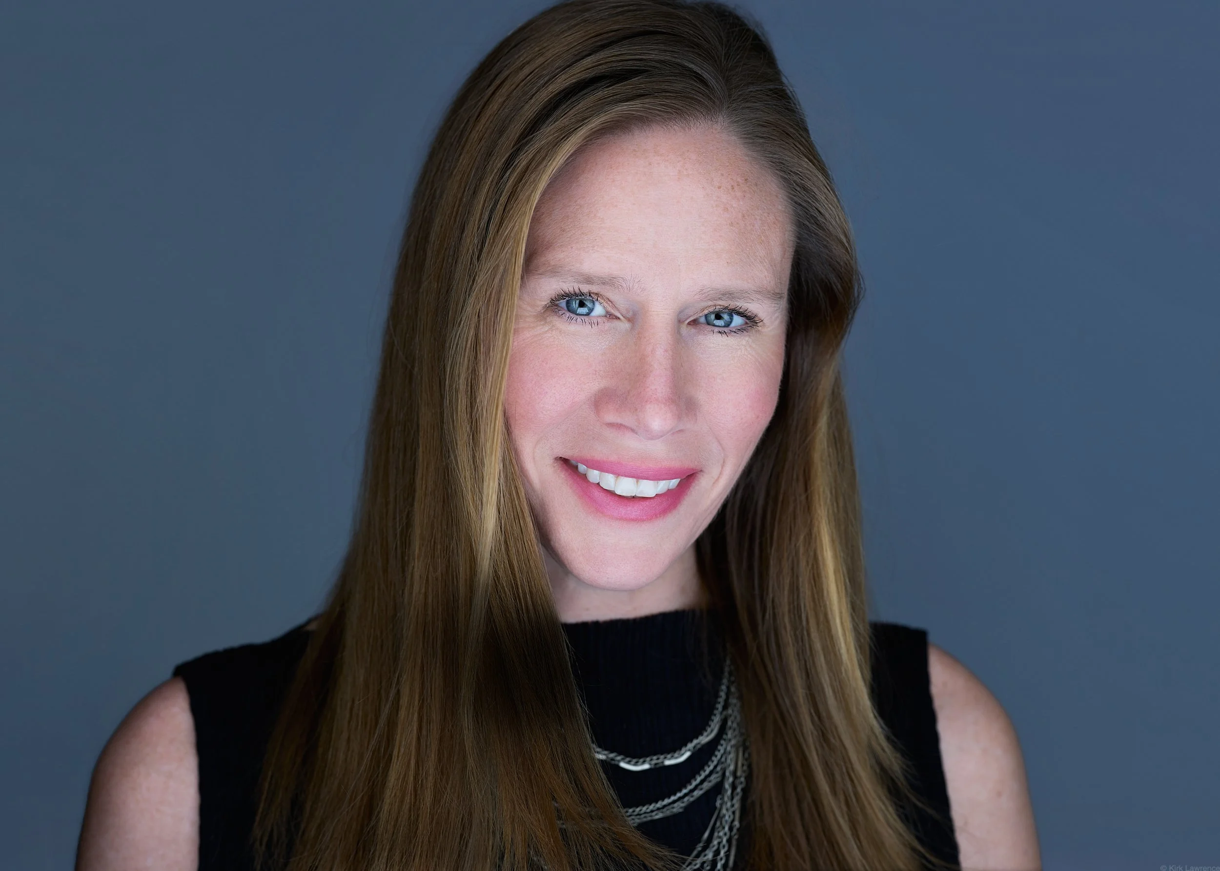 A woman with long red hair, green eyes, and a friendly smile, wearing a black sleeveless top and layered silver necklaces, stands against a gray background.