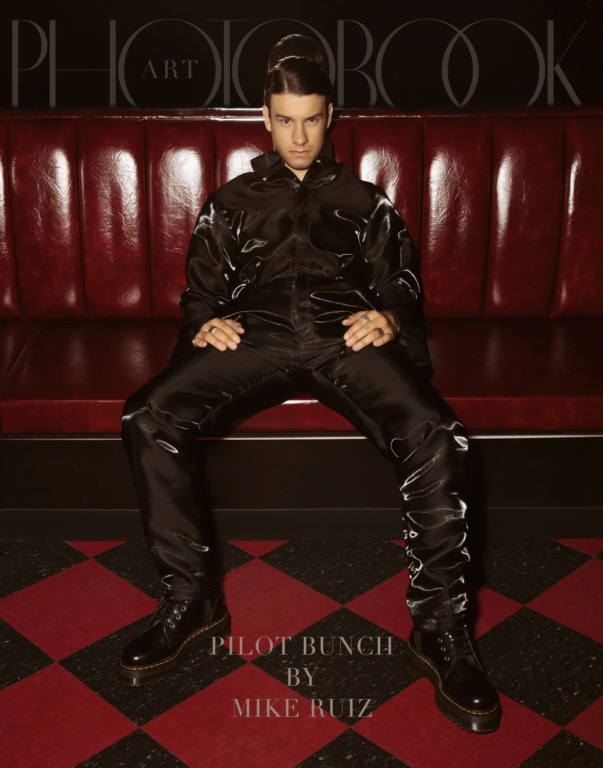 A portrait features a model sitting on a red leather booth, wearing a glossy black satin outfit with black boots.