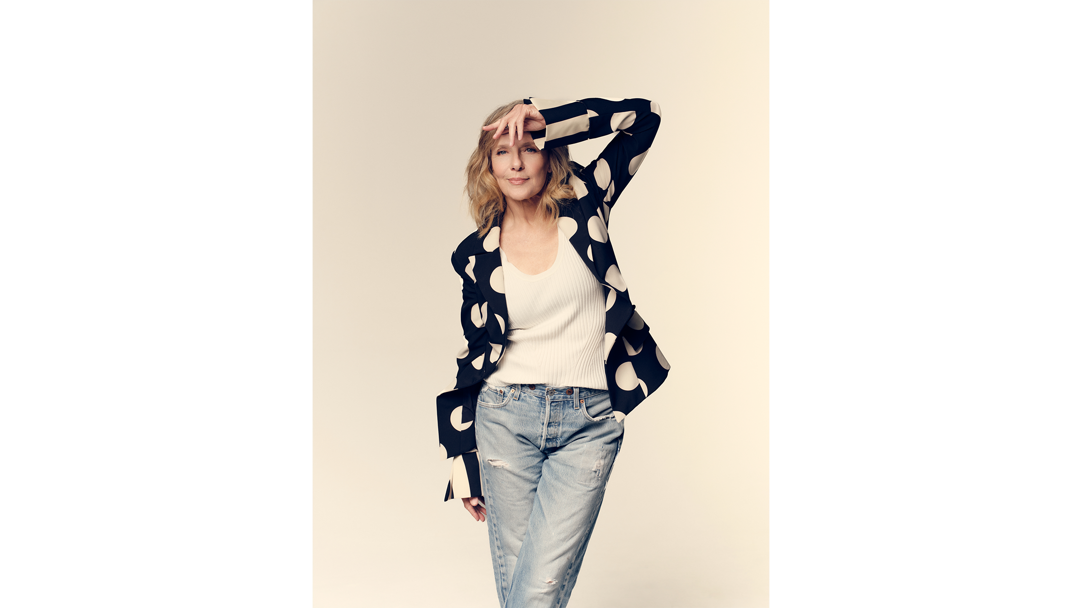 A portrait of Rosalee Mayeux posing with her hand on her forehead, wearing a black blazer with large white polka dots, a white ribbed tank top, and light-washed jeans.