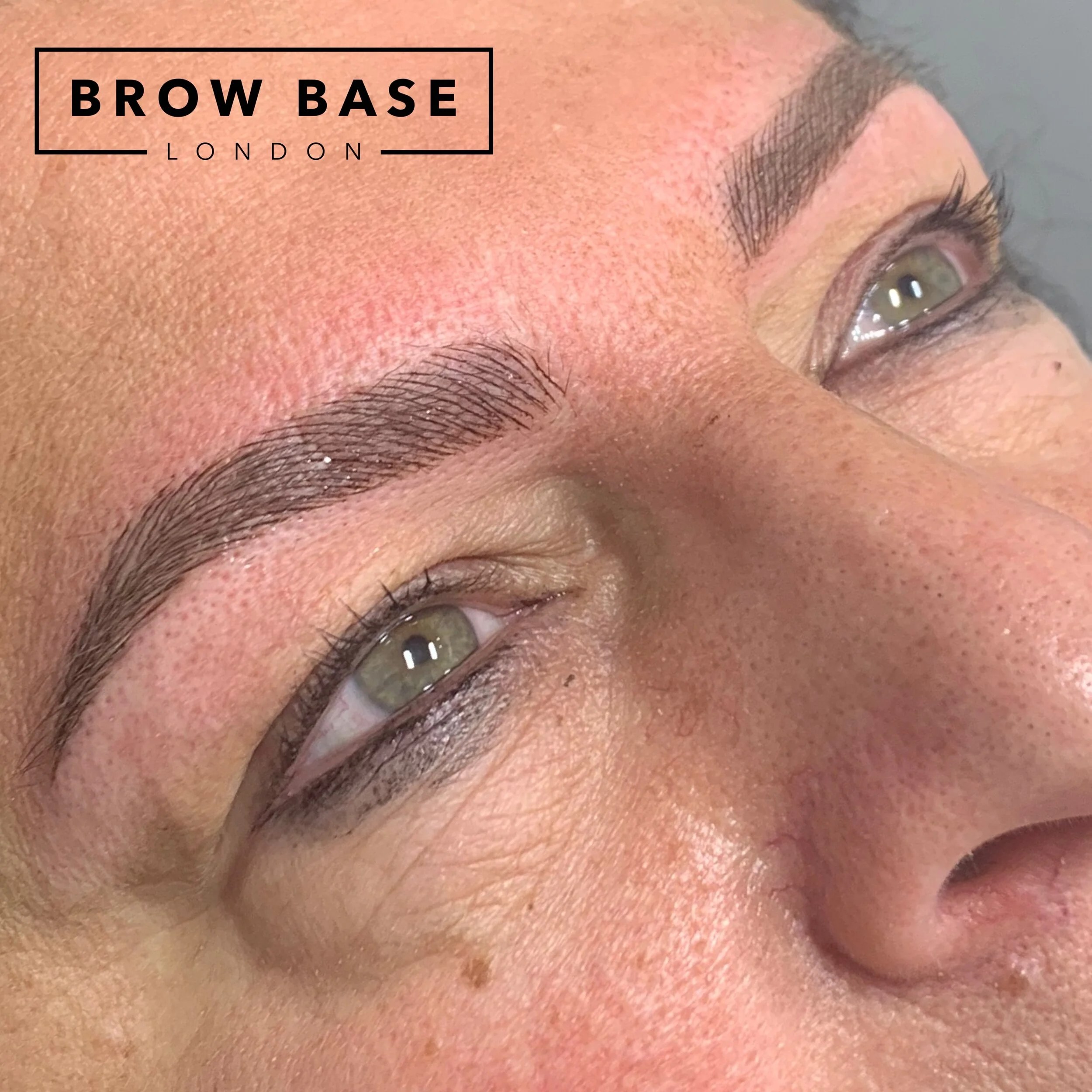 Close-up of a person's face focusing on freshly microbladed eyebrows with natural brow shape, and the logo of 'Brow Base London' in the top left corner.