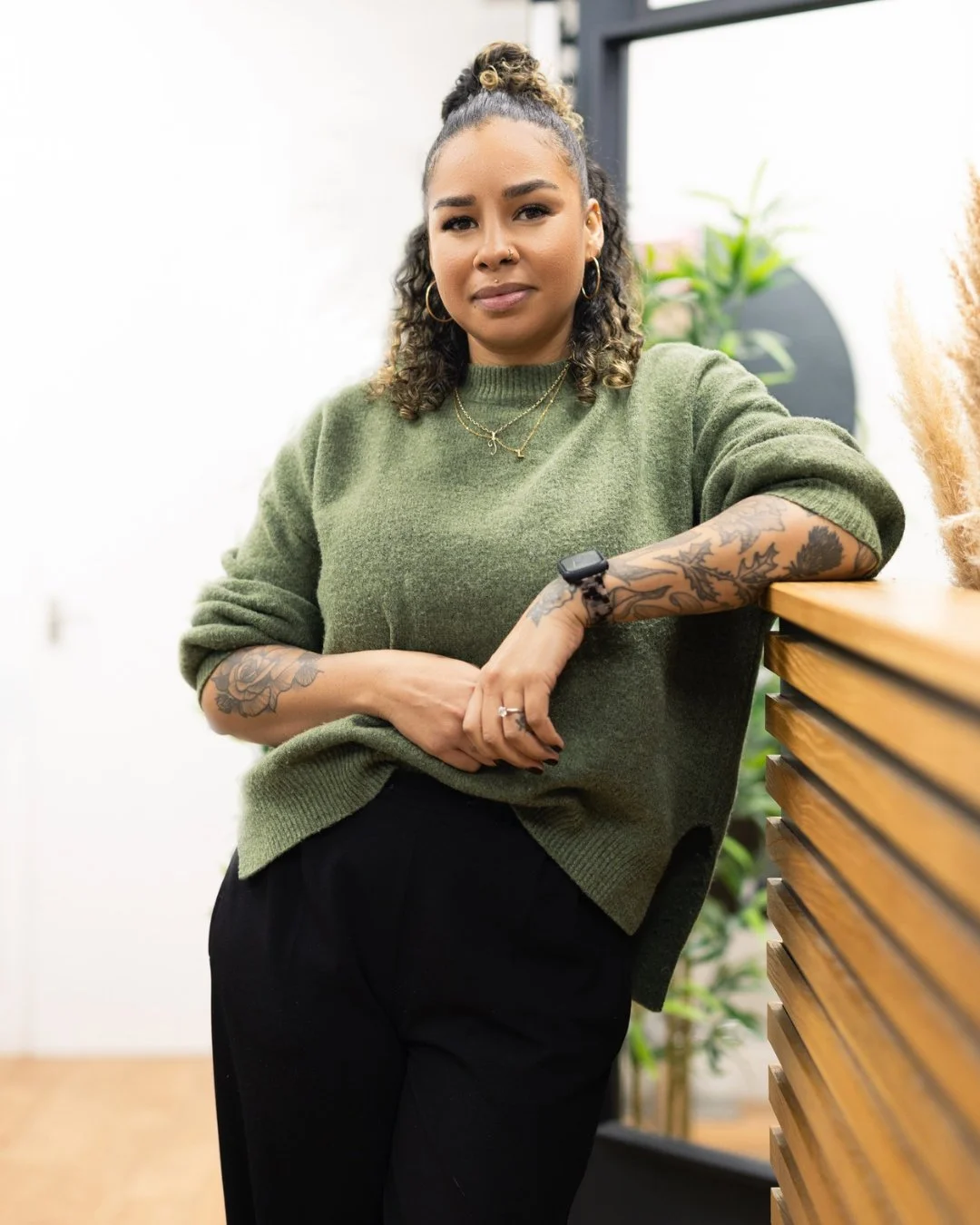 A woman with curly hair tied in a bun, wearing gold jewelry, a green sweater, and black pants, leaning on a wooden surface with her arms crossed, in a modern indoor space with plants.