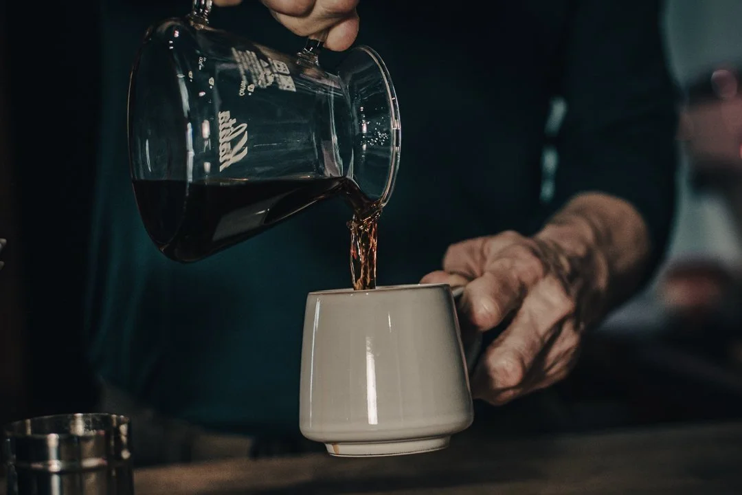 The owner of Trailhead pours hot delicious black coffee from a Kalita glass carafe into a beautiful white ceramic cup.