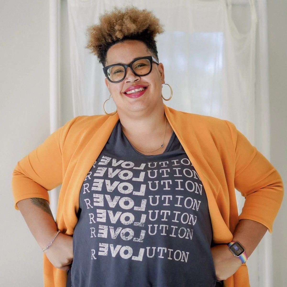 A smiling woman with glasses and large hoop earrings, wearing an orange blazer over a black T-shirt with the word 'REVOLUTION' printed multiple times, stands with her hands on her hips in front of a white curtain.