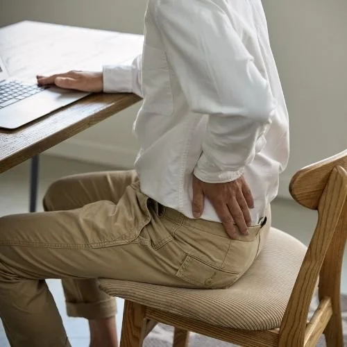 Man wearing khaki pants and a white shirt sitting on a chair in front of a desk with a laptop. His left hand is on his lower back as if he's in pain.