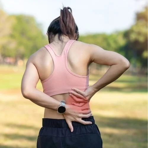 Woman with brown hair in a ponytail wearing a sports bra and black shorts. Her hands are behind her on her lower back to indicate pain in the region.